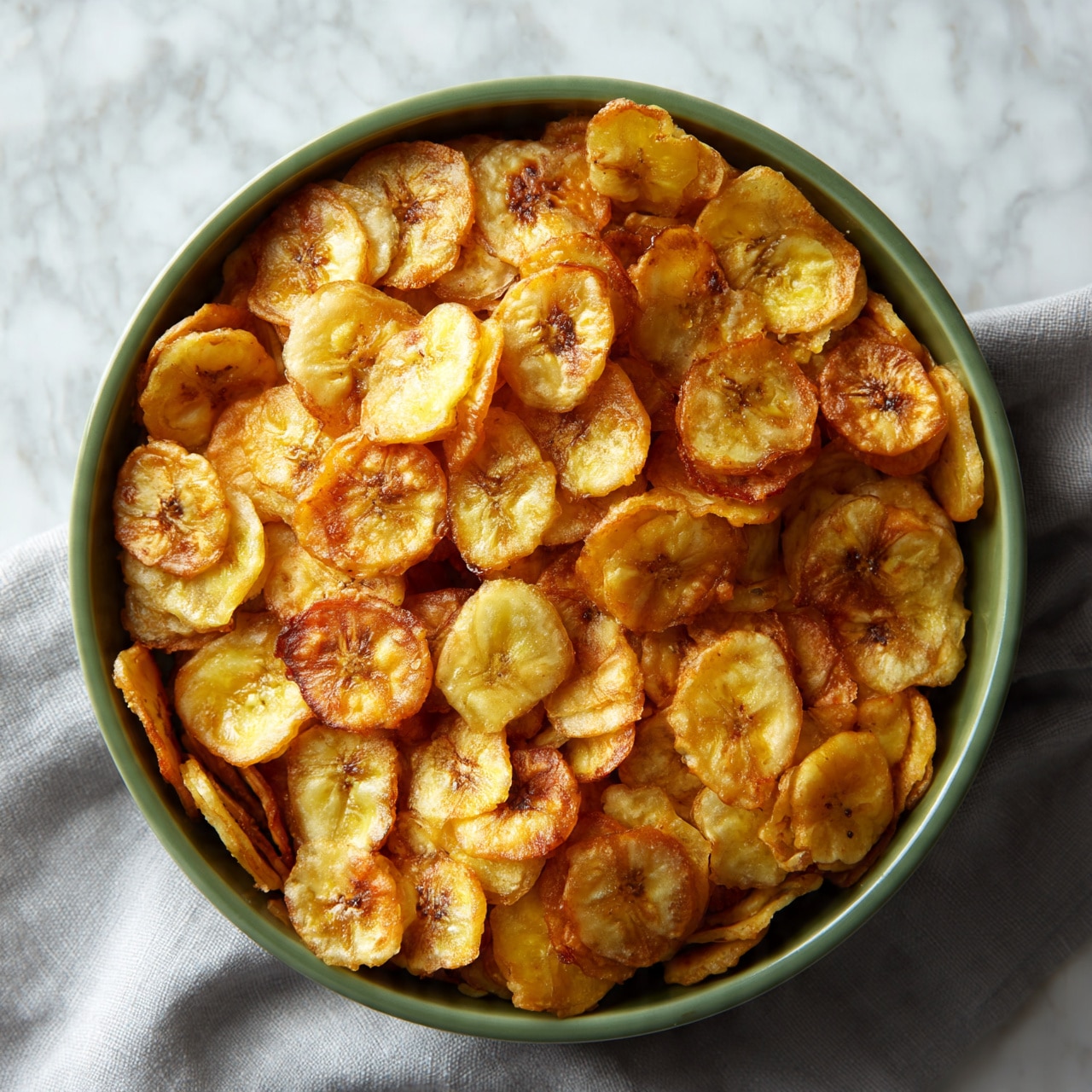 square image A green oval bowl is filled with one layer of round, thin, golden-brown banana chips. The chips have a slightly crispy texture with some darker spots, showing their fried nature. The bowl is placed on a soft grey cloth, but the background is changed to a white marbled texture. The banana chips cover the entire bowl evenly, showing some light yellow and caramel shades. Photo taken with an iphone --ar 4:5 --v 7