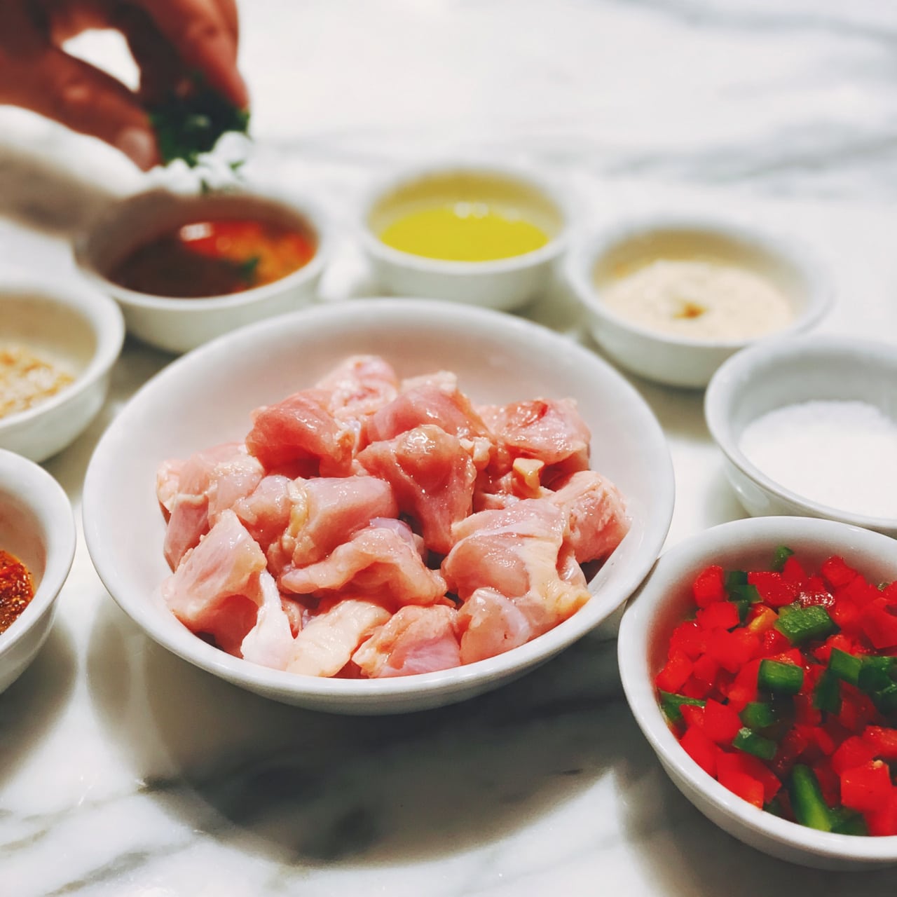 square image The image shows a white bowl in the center filled with pink raw chicken pieces. Around this bowl, there are several small white bowls arranged in a circle. One bowl contains bright red and green chopped bell peppers with a fresh texture. Other bowls hold different colored sauces and spices including a reddish-brown sauce, a yellow oil, a creamy white sauce, and white salt or powder. A woman's hand is seen at the top left holding some chopped green herbs over one of the bowls. The whole setup is placed on a white marbled surface that adds a clean and bright look to the image. photo taken with an iphone --ar 4:5 --v 7
