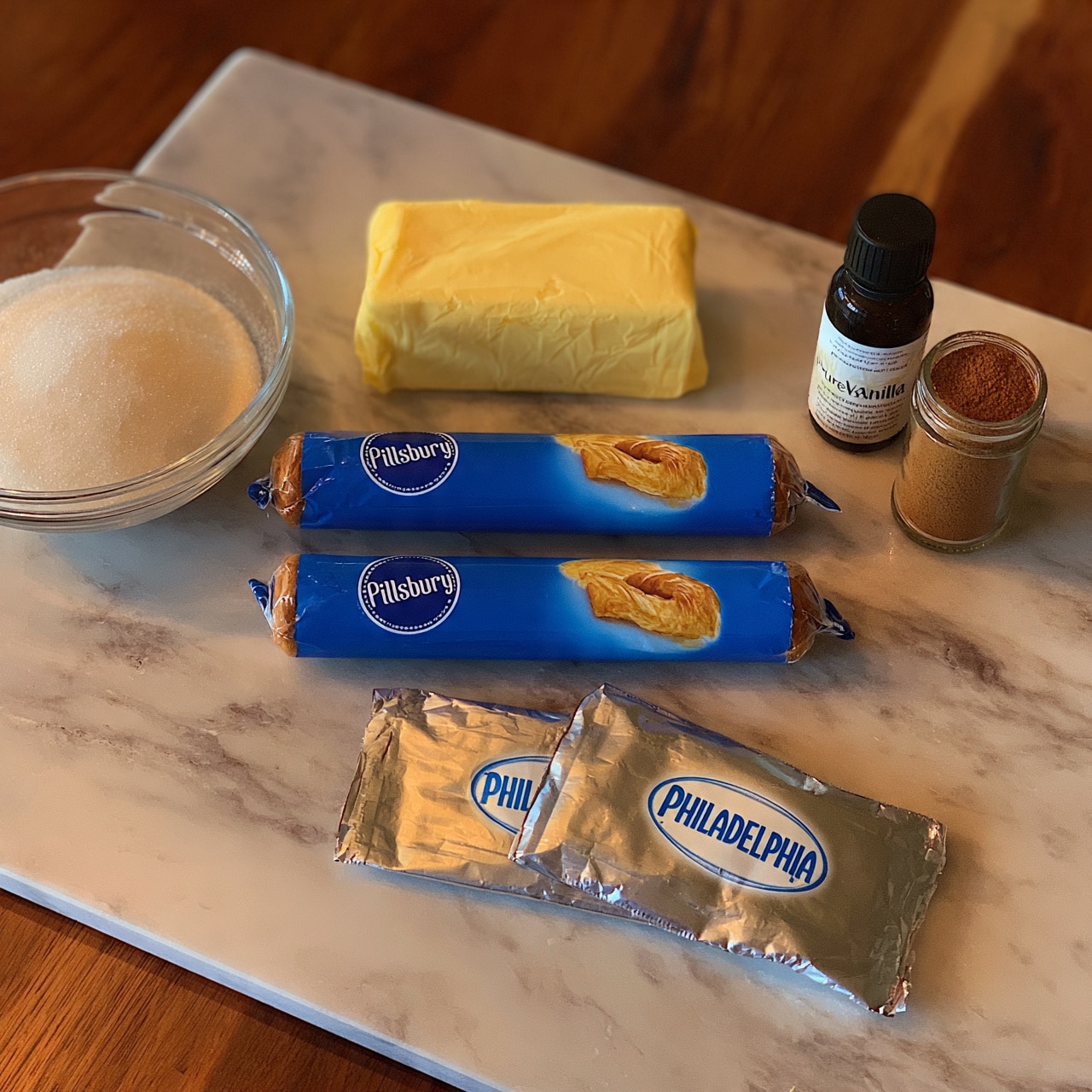 square image The image shows a wooden surface with baking ingredients neatly arranged. There are two blue tubes of Pillsbury Crescents aligned side by side in the center. Above them is a rectangular block of unsalted butter wrapped in yellow paper. To the right, there is a small dark bottle of pure vanilla extract standing next to a jar of ground cinnamon. Below the crescent tubes, two silver packages of Philadelphia cream cheese lie flat, and to the left, a clear glass bowl is filled with white granulated sugar. The scene is set on a white marbled texture surface. photo taken with an iphone --ar 4:5 --v 7