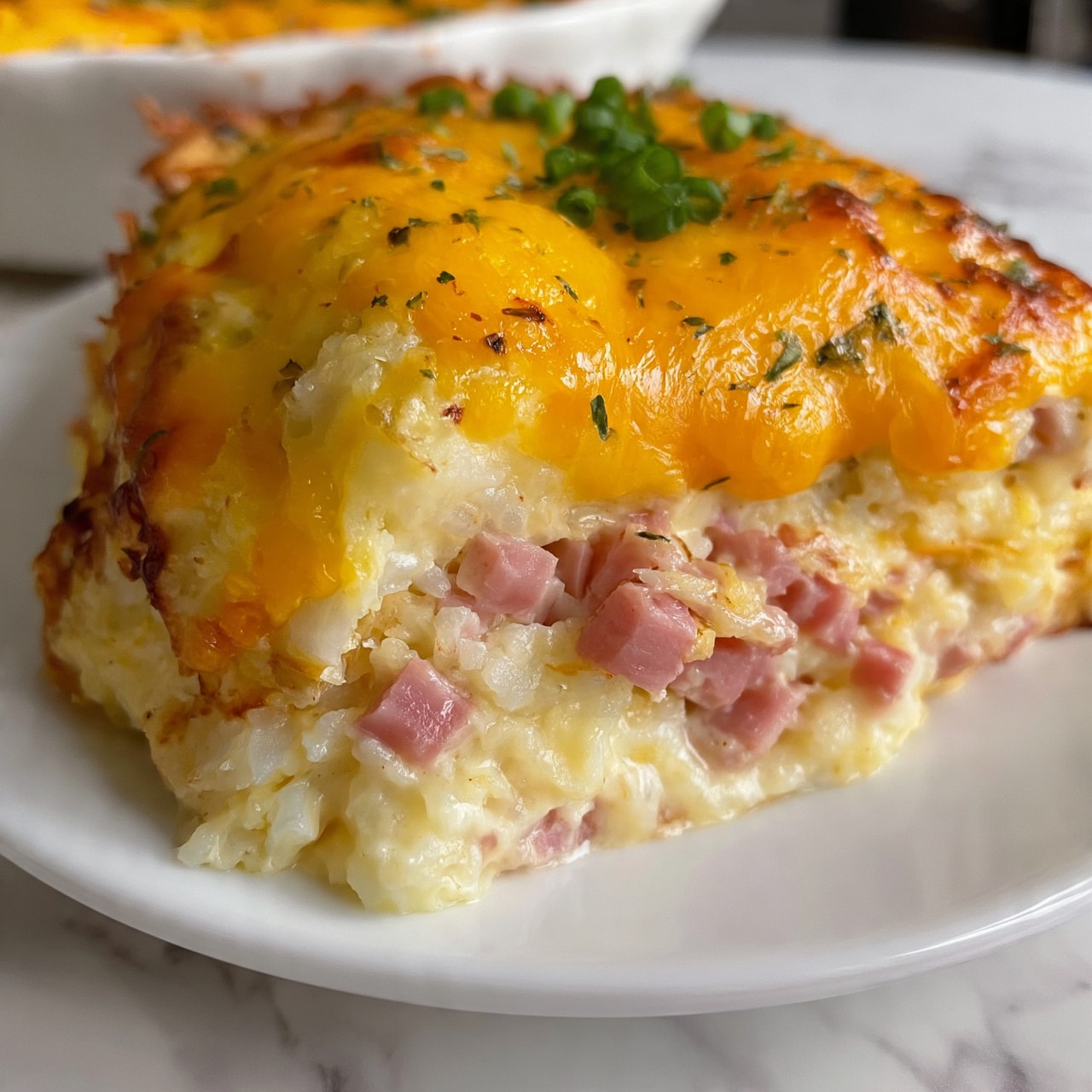 square imag  The dish shows a close-up of a baked casserole with at least three layers: the bottom layer is creamy and pale yellow with visible soft rice or potato texture, the middle layer consists of diced pink ham pieces mixed inside, and the top layer is melted bright yellow cheese, slightly browned in some spots, sprinkled with small bits of green herbs and chopped green onion. The food is on a white plate sitting on a white marbled surface. Photo taken with an iphone --ar 4:5 --v 7