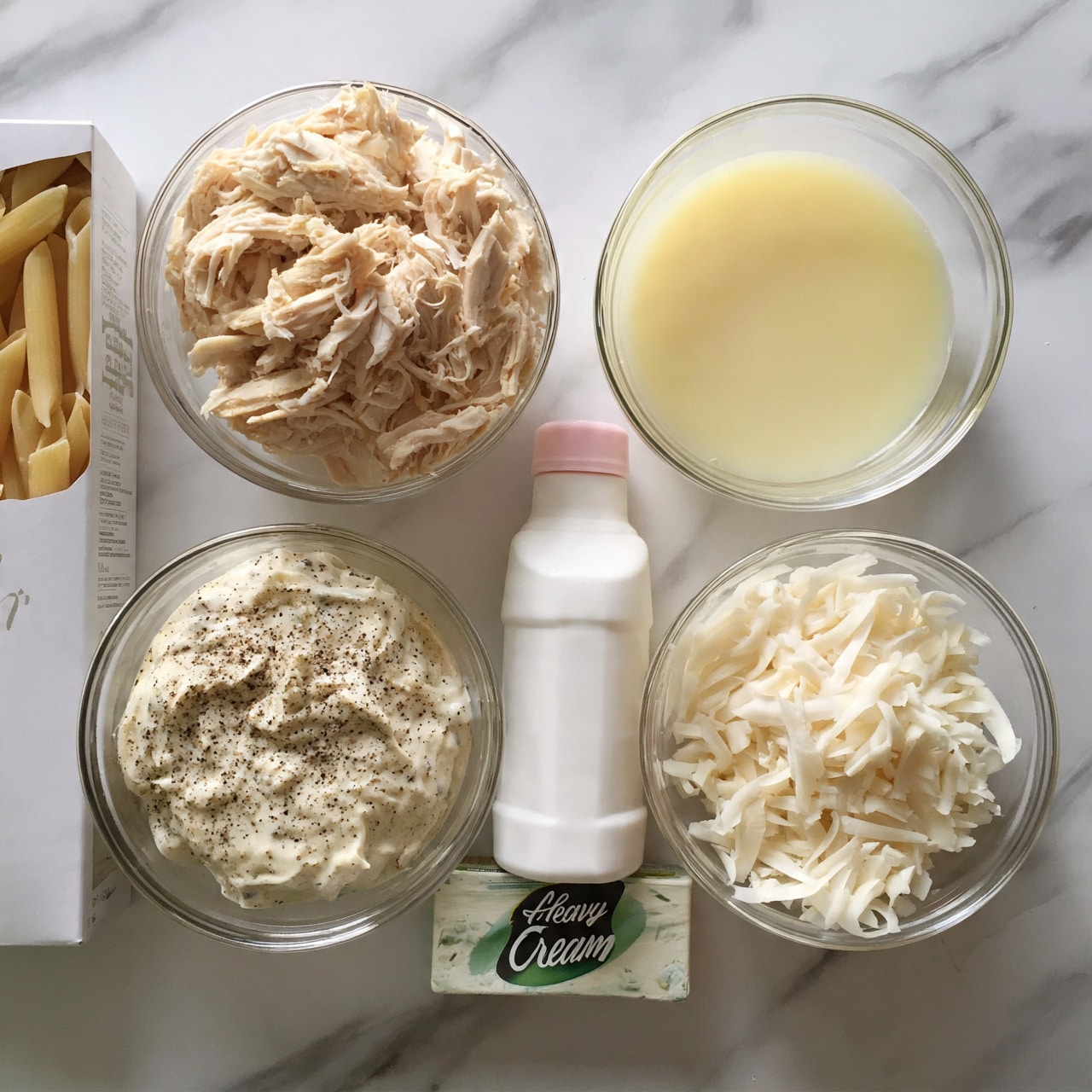 square image The image shows several ingredients arranged neatly on a white marbled surface. There is a white box of penne rigate pasta on the left side. To the right of the box, there are four clear glass bowls: one at the top filled with shredded light beige cooked chicken, one at the bottom left filled with a creamy mixture speckled with black pepper, one at the middle right filled with a light yellow liquid, and one at the bottom right filled with shredded white cheese. Between these bowls is a white plastic bottle with a pink cap labeled