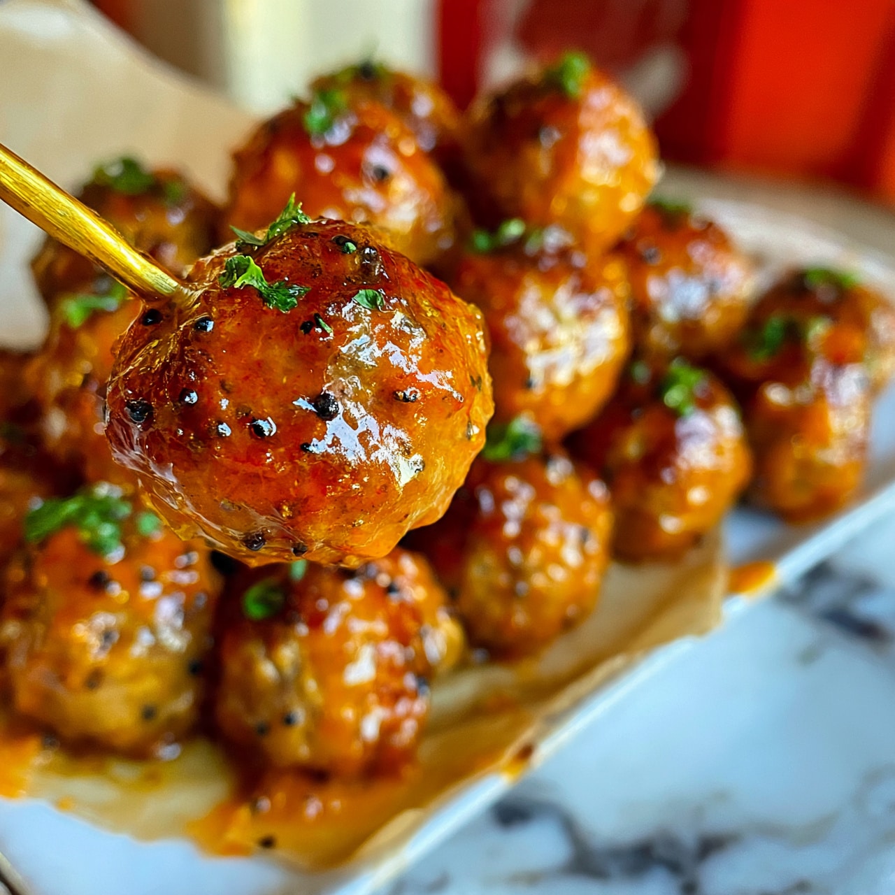 square image A group of small meatballs covered in shiny orange sauce with visible small black pepper bits, each meatball topped with bits of fresh green herbs, they sit tightly packed on a white marbled surface with some brown parchment paper underneath, one meatball is held up close on a golden skewer showing its glistening texture, the background is blurred with warm red and off-white colors. photo taken with an iphone --ar 4:5 --v 7
