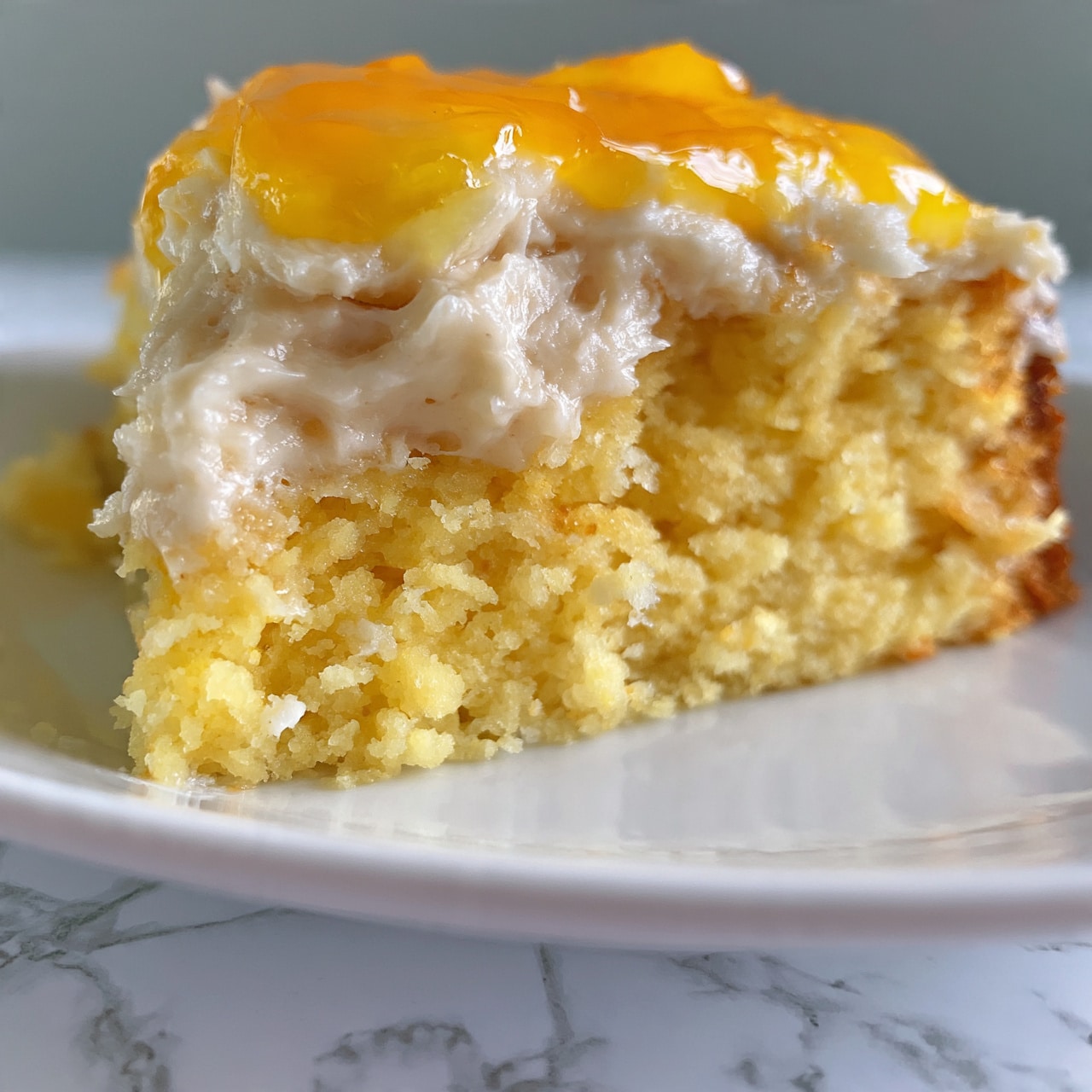 square image A close-up of a slice of yellow cake with a rough texture showing inside, topped with a creamy light beige frosting that looks soft and thick, and a bright orange-yellow layer of glaze or jelly on top, all resting on a white plate with a white marbled surface underneath. photo taken with an iphone --ar 4:5 --v 7