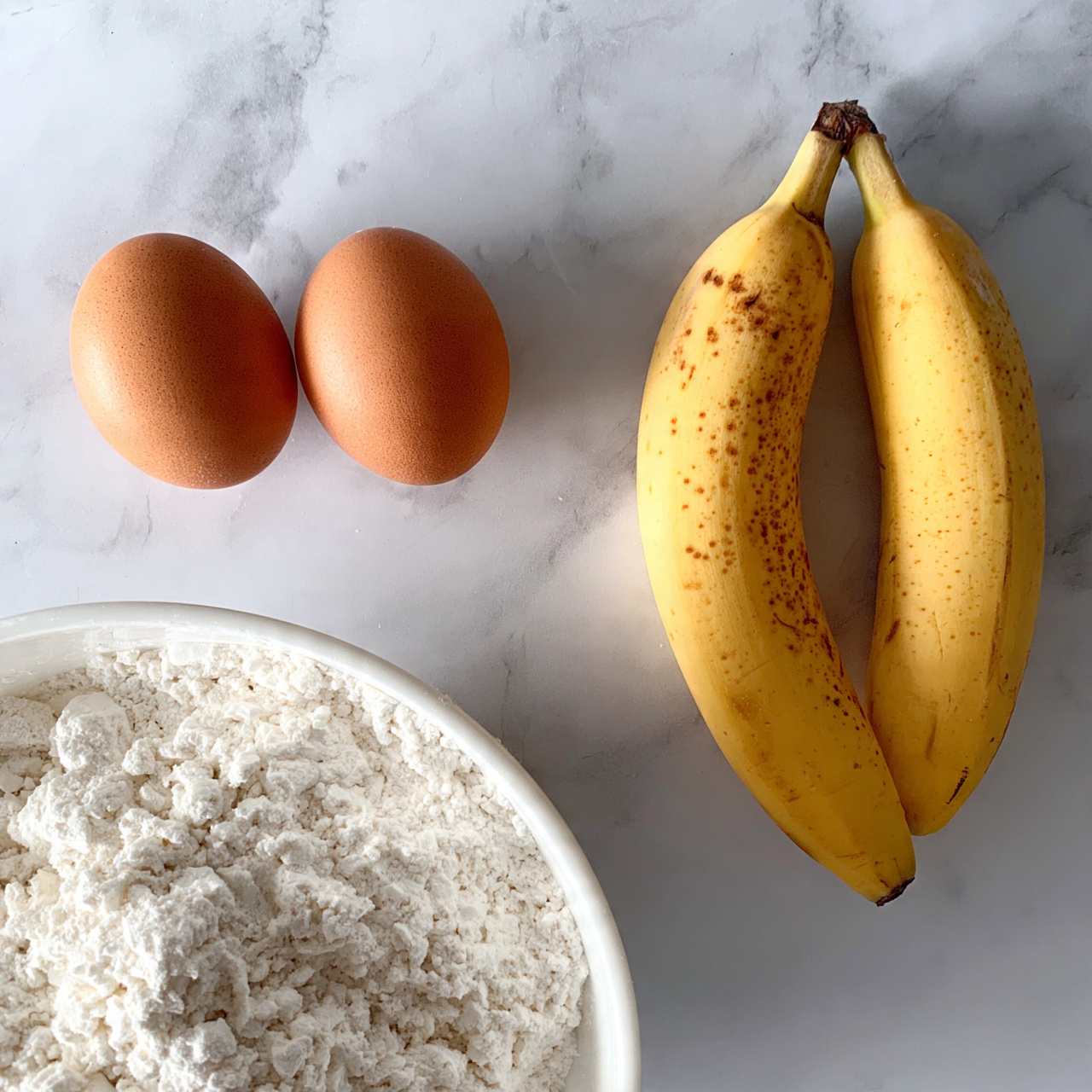 square image The image shows two brown eggs placed side by side on a white marbled surface on the left side. On the right side, there are two ripe yellow bananas with brown spots lying next to each other, also on the white marbled surface. Below the eggs, part of a white bowl filled with white flour is visible, with the flour having a soft, powdery texture. The overall scene is clean and simple with a focus on these four ingredients. photo taken with an iphone --ar 4:5 --v 7