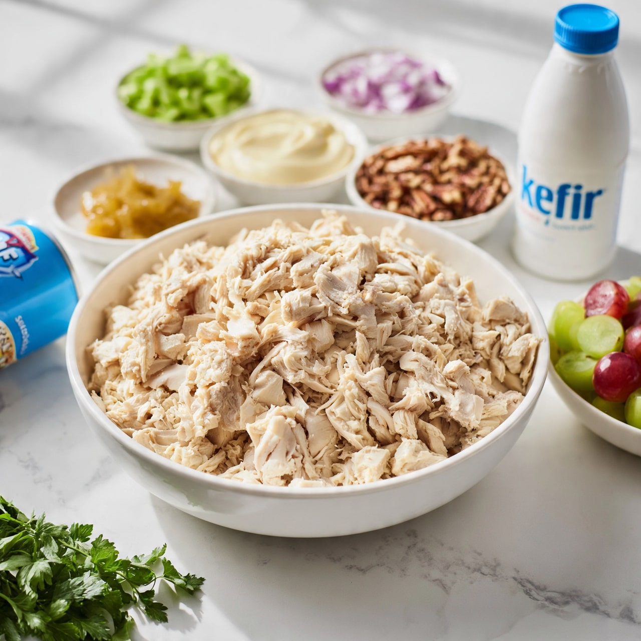 square image The image shows a large white bowl filled with shredded light-colored chicken placed in the center on a white marbled surface. Surrounding the bowl are smaller white bowls holding chopped green celery pieces, chopped red onion, mayonnaise, chopped pecans, sweet relish, a small amount of tan-colored sauce, and halved red and green grapes. A white bottle with blue cap labeled kefir stands on the right side, and a blue can of crescent rolls lies on the left. Fresh green parsley is positioned near the bottom left corner. The whole setup is clean and organized with soft natural lighting, photo taken with an iphone --ar 4:5 --v 7