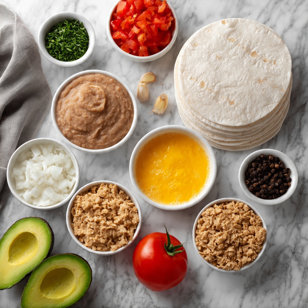 square image A white marbled surface with a neat arrangement of various ingredients. At the center right are stacked white tortillas. Surrounding them, there is a small white bowl with refried beans showing a smooth, creamy brown texture, another bowl with diced white onions, a larger bowl containing a bright yellow beaten egg mixture, and a bowl with light brown crumbled texture resembling cooked ground meat or textured soy. Fresh ingredients include two halves of a green avocado at the bottom left, a whole red tomato near the center, and a small pile of vibrant red diced tomatoes in a white bowl near the top center. Additional small white bowls hold finely chopped green herbs, whole black peppercorns, and a few garlic cloves. A gray cloth is partially visible on the left. Photo taken with an iphone --ar 4:5 --v 7