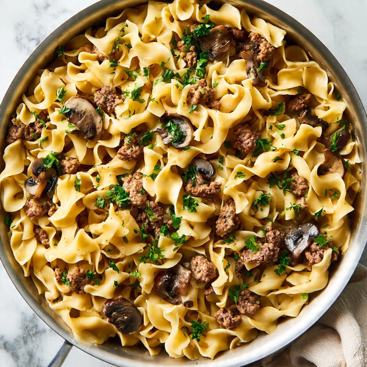 square image A large silver pan filled with three layers of wide, wavy egg noodles that are creamy light yellow. Mixed inside are small chunks of cooked ground meat in brown and pieces of sliced mushrooms in dark brown and gray. The dish is sprinkled with small bright green parsley leaves on top, giving fresh color contrast. The pan is on a white marbled surface with a beige cloth nearby. photo taken with an iphone --ar 4:5 --v 7