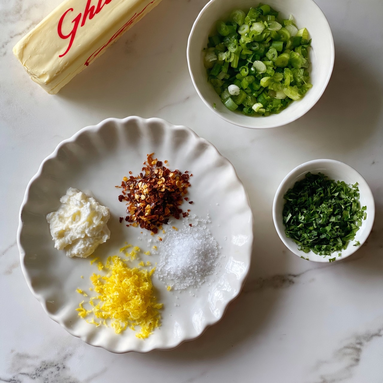 square image The image shows a white marbled surface with four white dishes arranged neatly. On the top left, there is a stick of butter with red lettering. To the right of the butter is a small white bowl filled with chopped green onions. Below them, there is a white scalloped plate holding four ingredients each in small piles: a white creamy substance to the left, bright yellow lemon zest at the bottom, crushed red pepper flakes at the top, and sea salt scattered across the plate. To the right of the scalloped plate is a tiny white bowl filled with finely chopped fresh green herbs. photo taken with an iphone --ar 4:5 --v 7