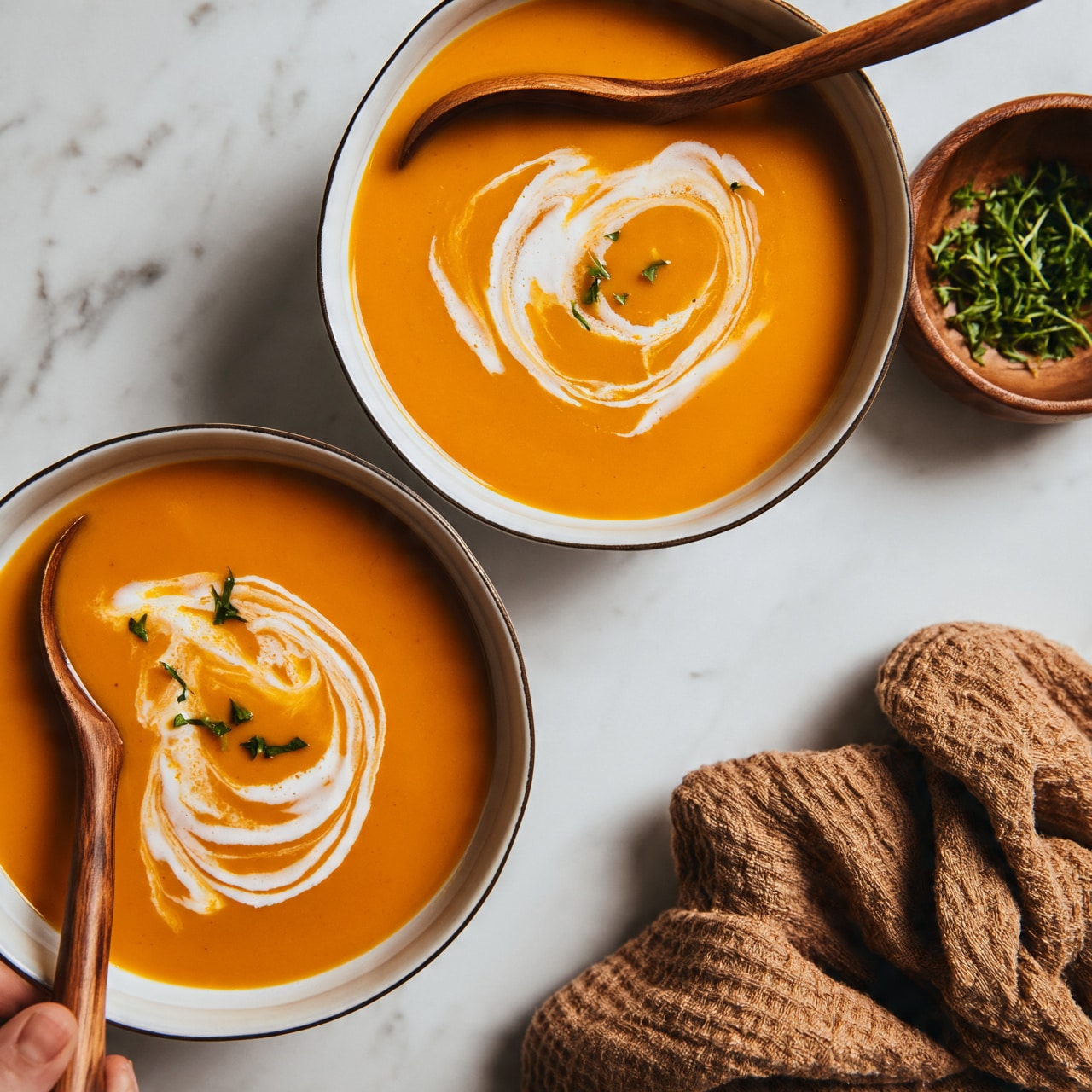 square image The image shows two bowls of smooth orange soup, each with a swirl of white cream and small pieces of green herbs on top. The bowls are white with a thin dark rim and sit on a white marbled surface. To the right, there is a small wooden bowl with more green herbs inside and a wooden spoon resting across the top bowl. A woman's hand holds a wooden-handled spoon on the left side near the bottom bowl. A textured brown cloth lies folded to the right below the bowls. photo taken with an iphone --ar 4:5 --v 7