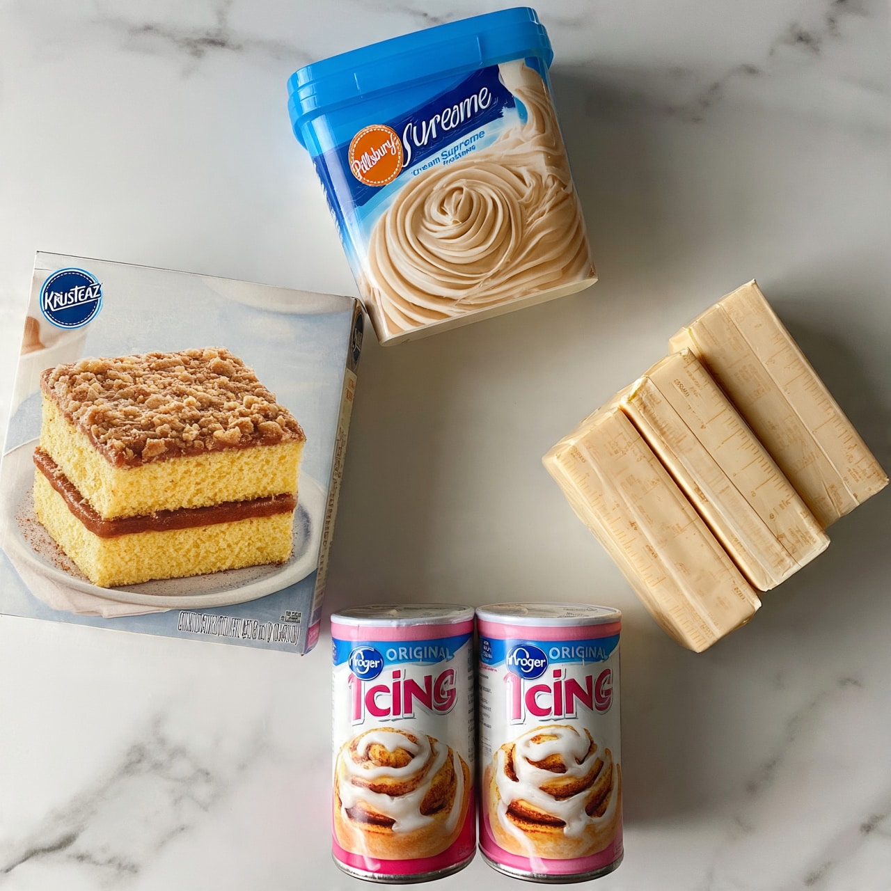square image The image shows a flat lay of baking ingredients on a white marbled surface. On the left side, there is a box of Krusteaz Cinnamon Swirl crumb cake and muffin mix with a picture of a square cake with two yellow cake layers and a thick brown cinnamon swirl in the middle and a crumb topping. Above it, there is a container of Pillsbury Creamy Supreme cream cheese frosting with a blue lid and swirled beige cream cheese frosting designed on the label. Below the box and frosting, two pink and white cans of Kroger Original Icing for cinnamon rolls are placed side by side, each showing an image of cinnamon rolls with white icing on top. On the right side, there are three sticks of butter in beige wrappers with measurement markings, arranged closely together. The whole setup looks clean and bright. photo taken with an iphone --ar 4:5 --v 7