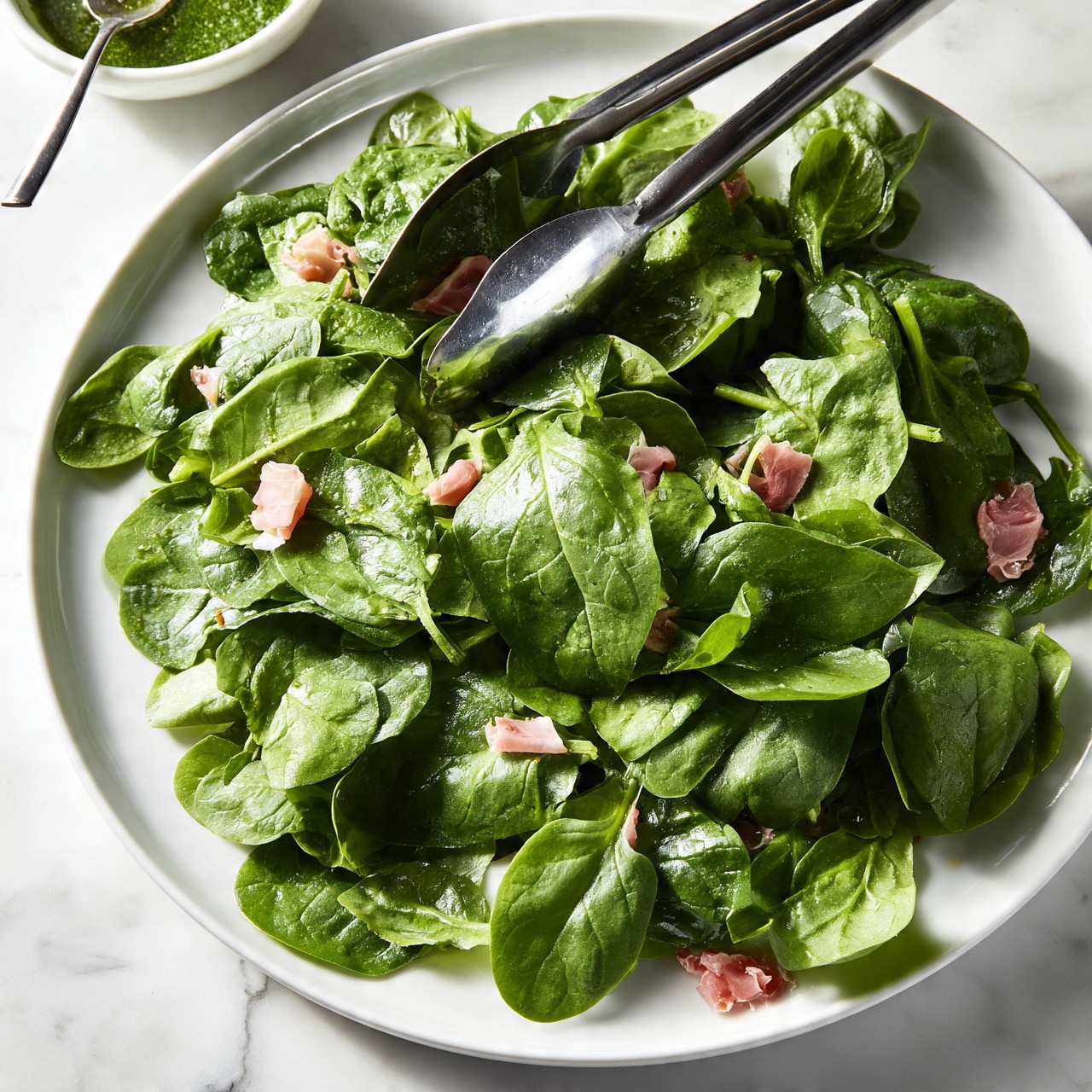 square image A large white plate is filled with fresh green spinach leaves, some with a smooth, slightly shiny texture, layered loosely across the plate. Among the spinach leaves are small pieces of light pink cured meat scattered unevenly. A pair of shiny metal tongs rests on top of the salad near the center. To the upper left of the plate, a small white bowl with a green sauce and a spoon sits on a white marbled surface. The lighting is bright and natural, emphasizing the freshness of the ingredients photo taken with an iphone --ar 4:5 --v 7