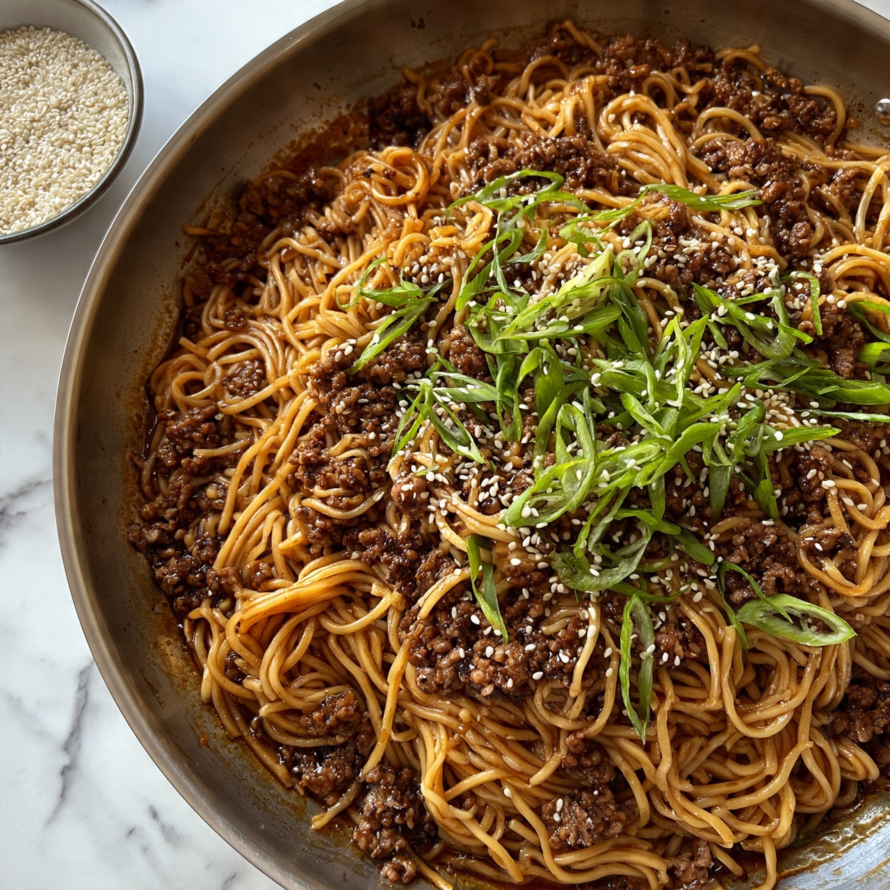 square image A close-up view of a pan filled with noodles mixed with ground meat, all coated in a shiny, dark brown sauce. The noodles are long and light brown, tangled with small pieces of darker brown cooked meat. On top, thin slices of green onion and white sesame seeds are scattered, adding a fresh green and white contrast to the dish. Next to the pan, there is a small bowl filled with white sesame seeds, all set on a white marbled surface. The metal pan shows a slight shine from the cooking oil. photo taken with an iphone --ar 4:5 --v 7