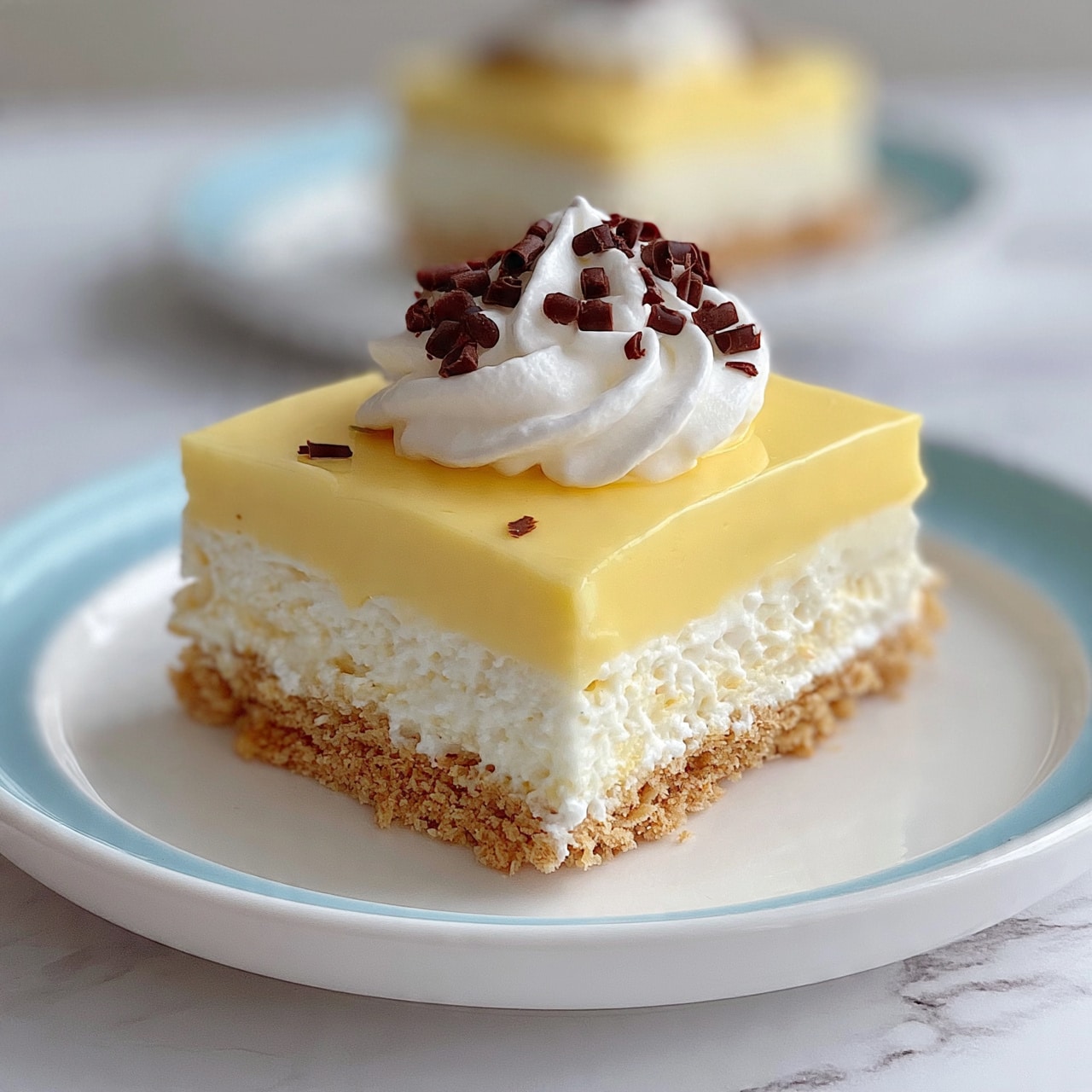 square image A slice of layered dessert sits on a white plate with a soft blue rim, placed on a white marbled surface. The bottom layer is crumbly and light brown, looking like crushed cookies or graham crackers. Above that is a thick, creamy white layer, topped by a smooth pale yellow layer that looks like custard or pudding. The top is finished with a fluffy white whipped cream dollop, decorated with small dark brown chocolate curls. Another slice of the same dessert is blurred in the background on a similar white plate with a blue rim. Photo taken with an iphone --ar 4:5 --v 7