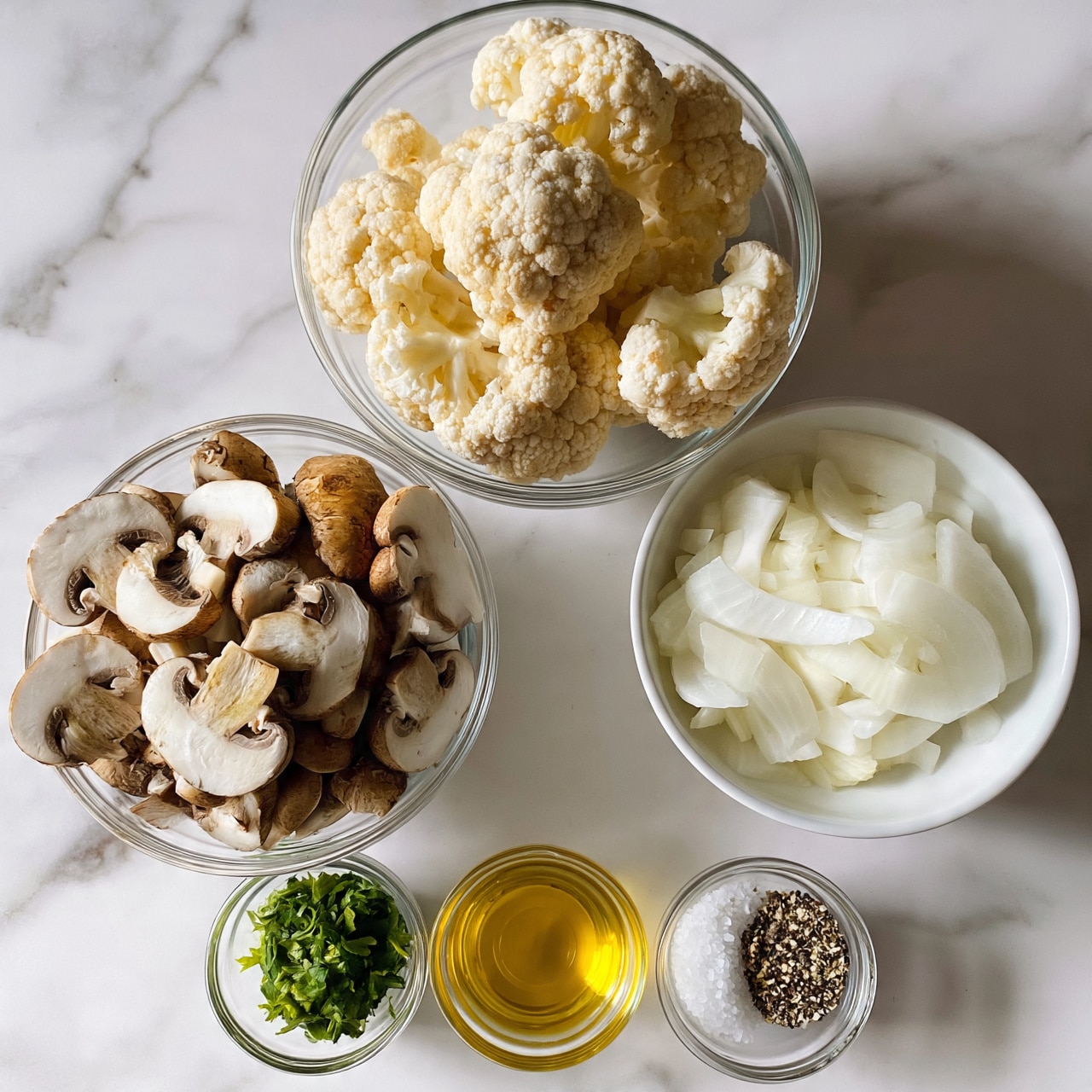  square image The image shows six clear glass bowls and one white bowl placed on a white marbled surface. The top clear bowl holds several pieces of pale cauliflower, with a textured and bumpy surface. Below it to the left is a clear bowl filled with halved mushrooms, with brown tops and white undersides. To the right of the mushrooms, a white bowl contains thinly sliced onion pieces that are translucent white with a smooth texture. In front of these, near the bottom, are smaller clear bowls arranged in a line: one with bright green fresh herbs, one containing golden yellow olive oil, and the last with coarse salt and cracked black pepper. The bowls are arranged neatly, showcasing the fresh ingredients in natural light, photo taken with an iphone --ar 4:5 --v 7