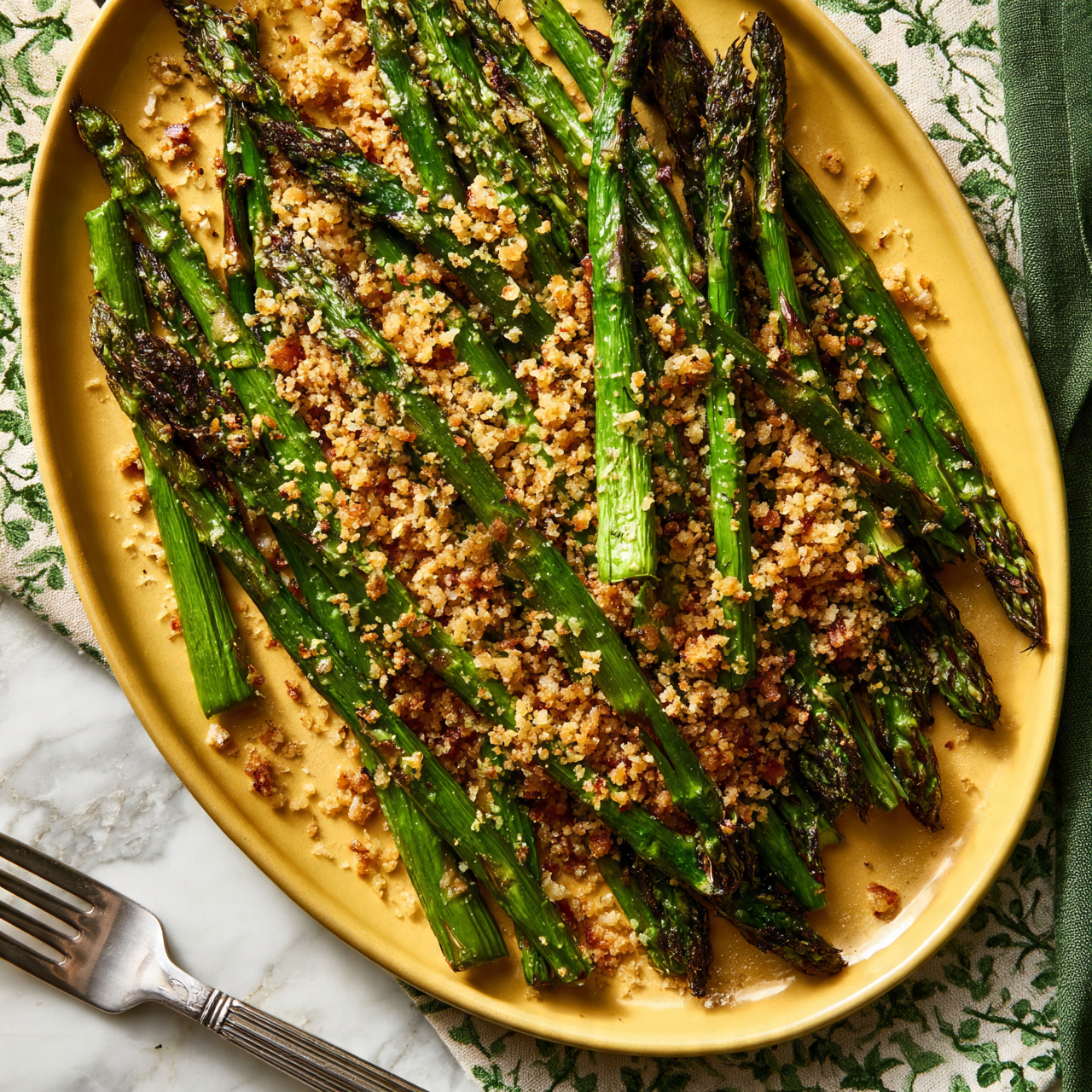 square image A yellow oval plate holds a pile of roasted green asparagus spears, each topped with a layer of crumbly, golden brown toasted breadcrumbs that add texture and small crunchy bits across the surface. The asparagus stalks are bright green with some darker charred spots from roasting. The plate rests on a white marbled surface partially covered by a fabric with a green vine pattern and a green napkin on the right side. A silver fork with a detailed handle is positioned at the bottom left corner of the image photo taken with an iphone --ar 4:5 --v 7
