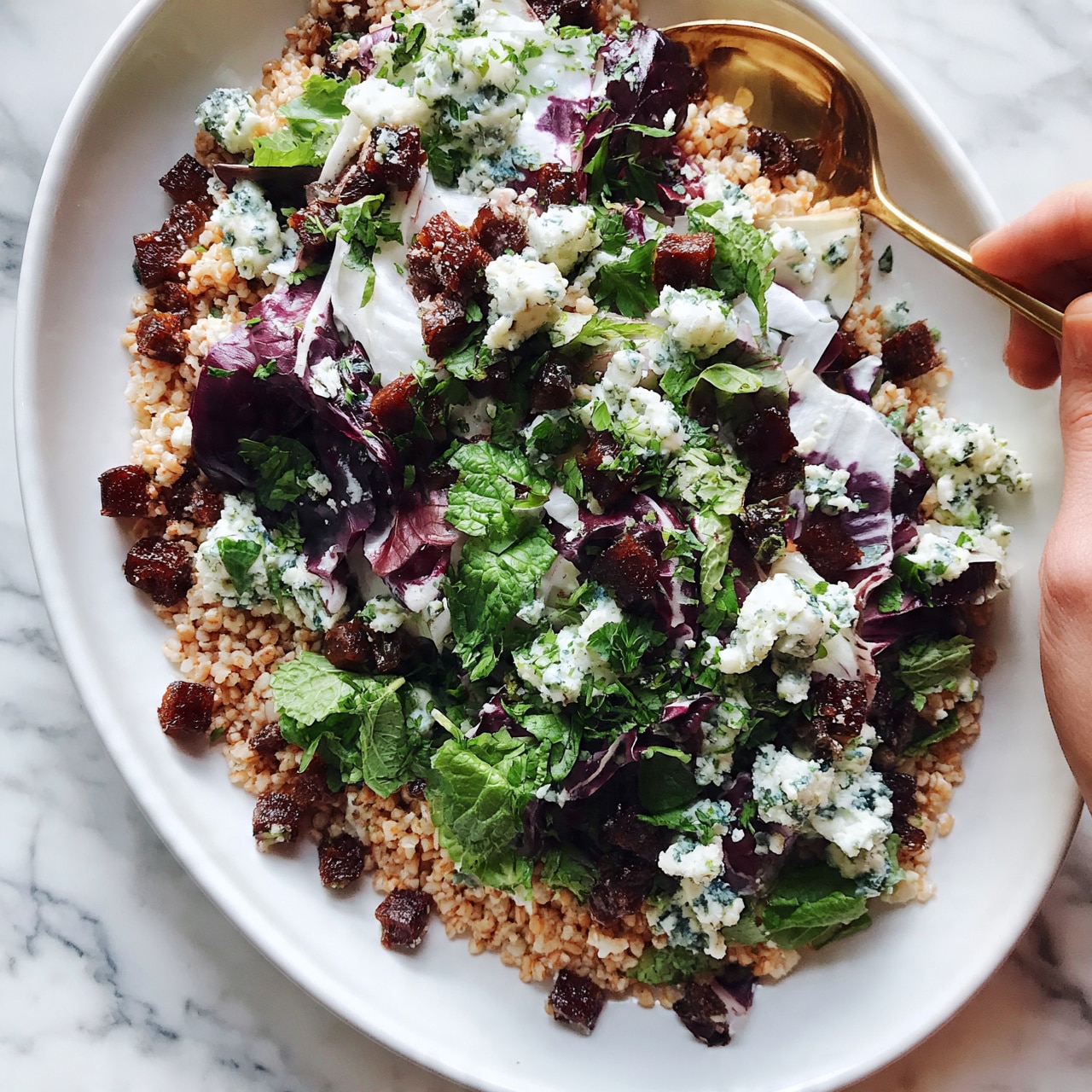 square image A white oval plate filled with a colorful salad featuring several layers: the base layer is light brown grains, scattered throughout are dark brown small cubes, likely dried fruit. Bright green leafy herbs are sprinkled all over the salad, mixed with deep purple and white leaves, and topped with small white chunks with blue-green specks, resembling cheese. The plate sits on a white marbled surface with a woman's hand holding a gold spoon partially visible on the right side. The lighting makes the salad look fresh and vibrant, highlighting the various textures and colors. photo taken with an iphone --ar 4:5 --v 7