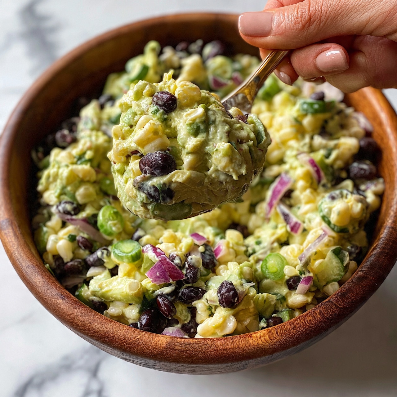 square image A close-up view shows a wooden bowl filled with a creamy salad. The salad has several visible layers including light green avocado, dark black beans, white corn kernels, and thin slices of purple-red onion scattered throughout. There are also small round green cucumber slices mixed in. A woman's hand holds a spoon lifting a large scoop of the salad, showing the creamy texture with fresh, colorful vegetables. The bowl sits on a white marbled surface photo taken with an iphone --ar 4:5 --v 7