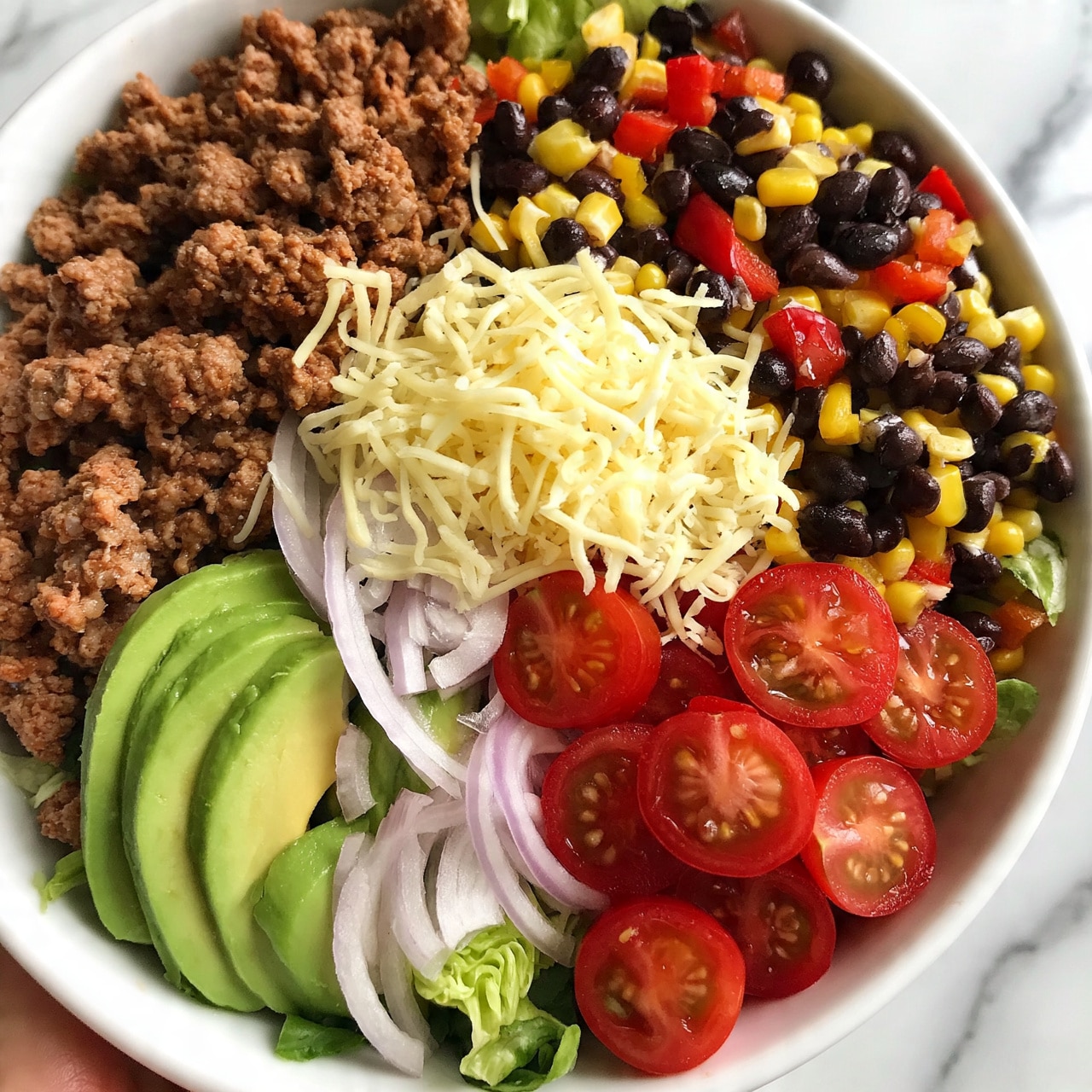 square image A white bowl filled with a colorful salad divided into different sections; one section has cooked ground meat with a brown, crumbly texture, another with sliced green avocado, thinly sliced light purple onions, and fresh cherry tomatoes cut in half showing bright red color; next to it is a mix of black beans, corn, and diced red peppers with yellow and black tones, topped with shredded pale yellow cheese, and a few green lettuce leaves peeking through. The bowl sits on a white marbled surface, with part of a woman's hand holding the bowl visible at the edge. photo taken with an iphone --ar 4:5 --v 7
