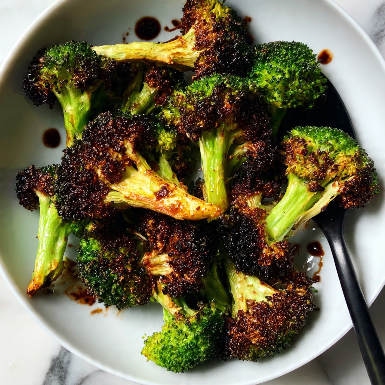 square image The dish shows many pieces of roasted broccoli with dark brown, almost black edges and bright green parts, placed in a white shallow bowl. The broccoli pieces have thick light green stems and textured tops that look crispy. Some drops of dark sauce are scattered around on the broccoli and bowl. A black spoon is under some broccoli on the right side. The bowl is set on a white marbled surface. Photo taken with an iphone --ar 4:5 --v 7