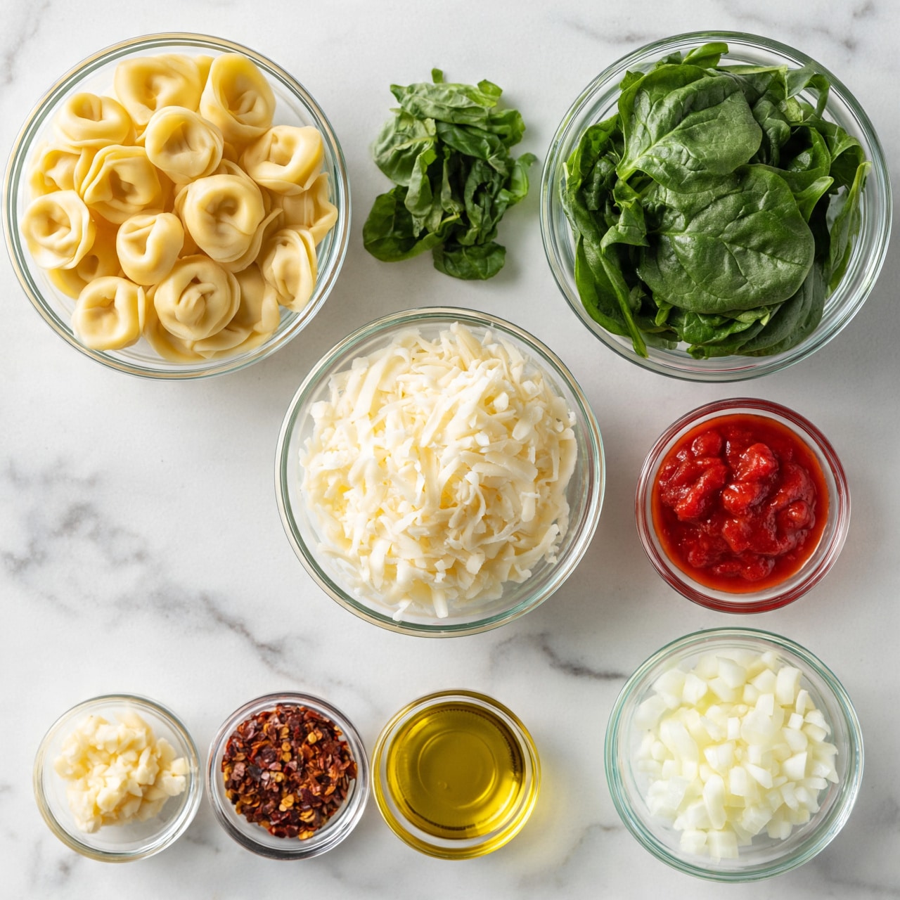 square image The image shows several clear glass bowls and small containers arranged on a white marbled surface. In the top row from left to right, there is a bowl filled with uncooked tortellini pasta that is pale yellow, a small pile of fresh green basil leaves, and a larger bowl filled with fresh spinach leaves that are dark green and leafy. Below, there is a bowl filled with shredded white cheese placed in the center. To the right of it is a small clear bowl with red tomato sauce. On the bottom left, there are four small containers holding minced garlic, red chili flakes, golden olive oil, and finely chopped white onions from left to right. A woman's hand is not visible but implied for arranging these elements. The lighting is bright and the setting appears clean and organized. Photo taken with an iphone --ar 4:5 --v 7