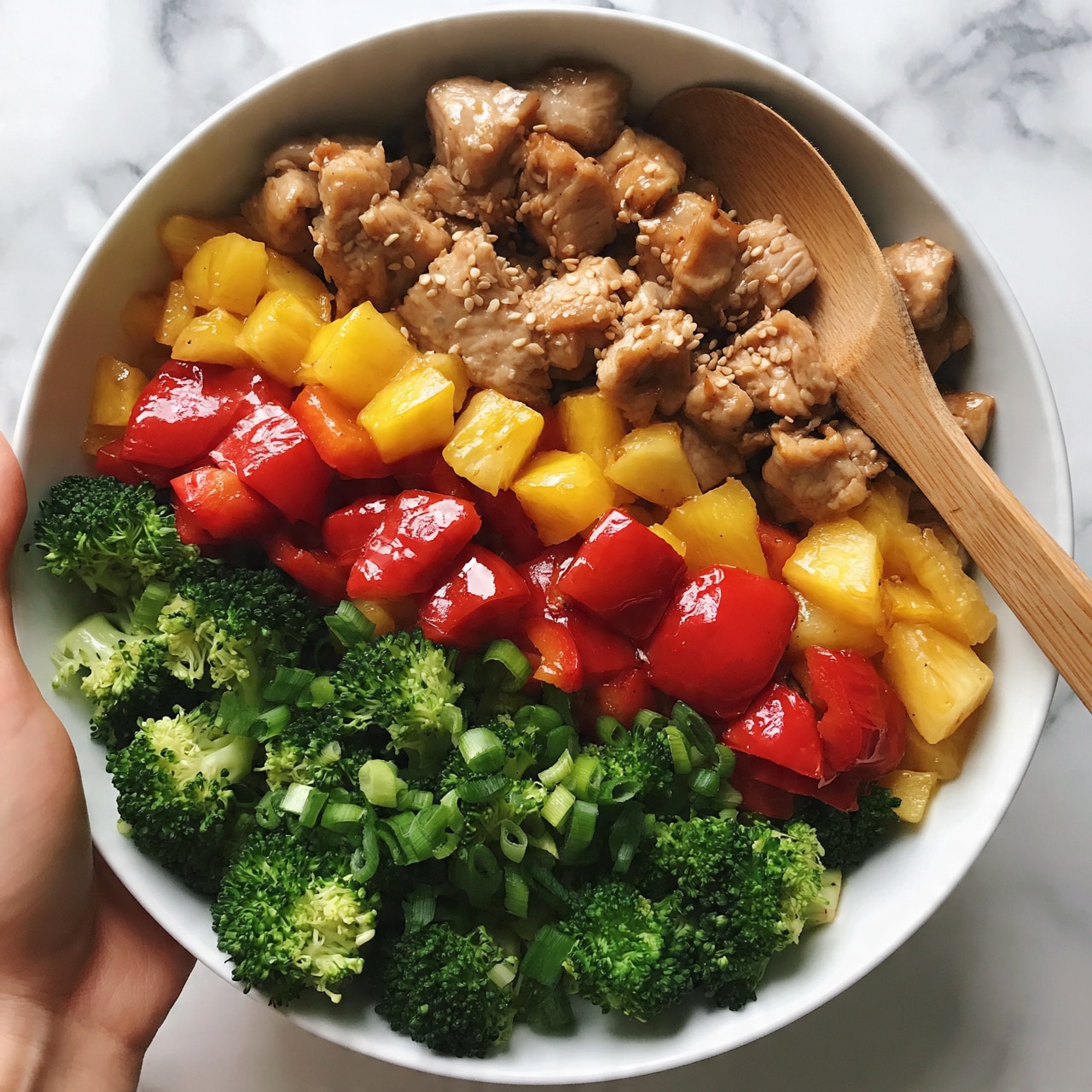 square image A white bowl on a white marbled surface filled with a colorful stir-fry dish showing three main layers: the first layer contains bright green broccoli florets and chopped green onions; the second layer has large, shiny chunks of red bell pepper and yellow pineapple pieces; the third layer consists of tender, light brown cooked chicken pieces mixed with sesame seeds scattered on top; a wooden spoon is placed on the right side of the bowl gently touching the food, and a woman's hand is holding the bowl from the left side photo taken with an iphone --ar 4:5 --v 7