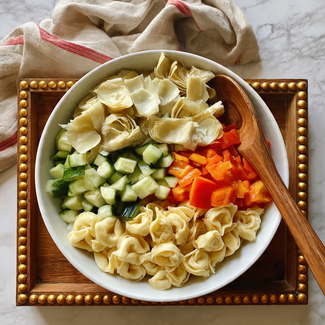 square image A white bowl filled with three main layers of food: the bottom layer is light yellow tortellini pasta, the middle layer has bright green cucumber chunks and orange-red bell pepper pieces, and the top layer shows pale, slightly translucent artichoke hearts scattered throughout. A wooden spoon with a smooth handle is resting in the bowl, partly submerged in the pasta mix. The bowl sits on a wooden tray with golden bead details on the edges, and a beige cloth with red stripes is casually draped in the background against a white marbled surface. photo taken with an iphone --ar 4:5 --v 7