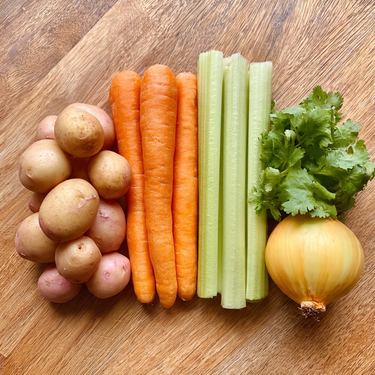square image A group of fresh vegetables is neatly arranged on a wooden surface with a light brown color and natural grain texture. On the left side, there are eight small round potatoes with smooth, light brown and pinkish skins, stacked in a loose cluster. To the right of the potatoes, three bright orange carrots with smooth skin are placed side by side vertically. Next to the carrots, three sticks of pale green celery with a firm, slightly ribbed texture lie parallel. On the far right, a small bunch of bright green cilantro leaves with jagged edges sits below a single round, yellow onion with a papery skin and a small root tip. photo taken with an iphone --ar 4:5 --v 7