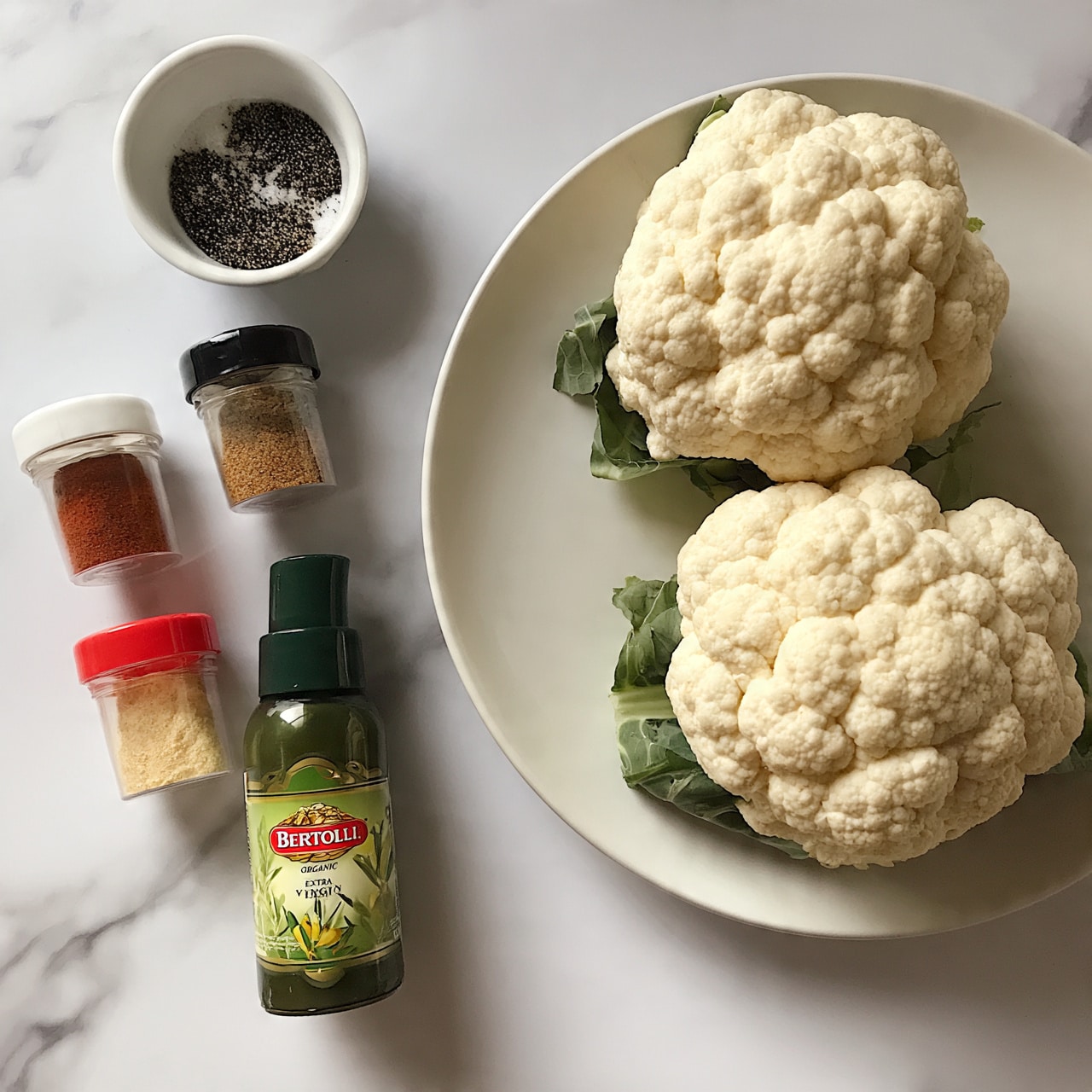 square image Two whole white cauliflowers with green leaves underneath, placed side by side on a large white plate, are positioned on the right side of the image. On the top left, there is a small white bowl with black salt and pepper powders inside. Below the bowl, there are four spice containers standing side by side: ground paprika in a clear container with a white lid, ground coriander in a taller clear container with a black lid, garlic powder in a short clear container with a red lid, and a green spray can of Bertolli organic extra virgin olive oil in front. All items are set on a white marbled surface. Photo taken with an iphone --ar 4:5 --v 7