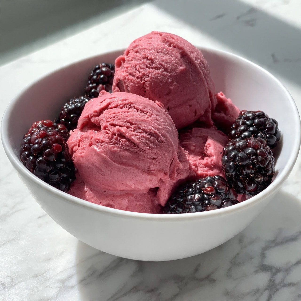 square image The image shows a white bowl filled with three scoops of dark pink ice cream that looks smooth with a slight texture on the surface. Around the ice cream, there are several fresh blackberries with a shiny, bumpy texture. The bowl sits on a white marbled surface. The photo is close-up, capturing the details of the ice cream and berries clearly. photo taken with an iphone --ar 4:5 --v 7