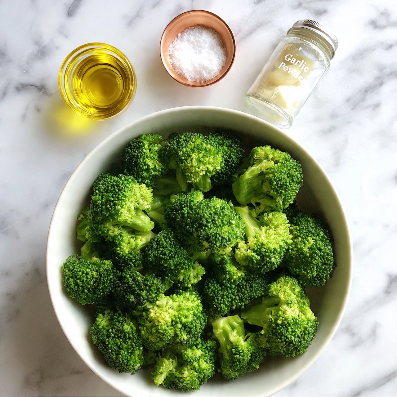 square image The image shows a white bowl filled with small green broccoli florets, each piece with a textured, bumpy surface and vibrant color. The broccoli is fresh and takes up most of the bowl. Above the bowl, there are three small round containers placed on a white marbled surface: on the left is a container with yellow oil, in the middle a copper bowl with white salt, and on the right a glass jar labeled