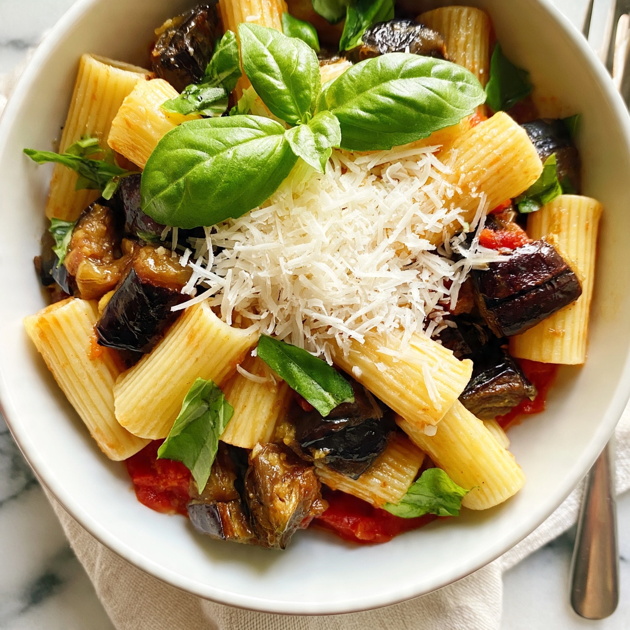 Ziti alla Norma (Eggplant and Tomato Pasta) Recipe 5 square image A white bowl filled with rigatoni pasta mixed with chunks of dark brown roasted eggplant and red tomato sauce at the bottom layer. On top, fresh green basil leaves are scattered, and a layer of finely shredded white cheese covers the center. The bowl is placed on a light cloth over a white marbled surface, with a metal fork nearby. photo taken with an iphone --ar 4:5 --v 7