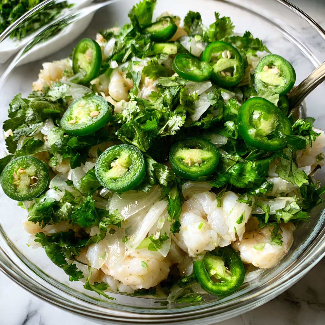 Grilled Avocado Halves Filled with Fresh Veggie Ceviche Recipe 4 square image A clear glass bowl filled with fresh shrimp salad showing three main layers: the base layer is white and firm shrimp pieces mixed with thin slices of translucent white onions, topped with vibrant green cilantro leaves and sliced bright green jalapeño peppers scattered evenly throughout. The ingredients look fresh and moist, with a spoon visible on the right side, resting inside the bowl. The bowl is placed on a white marbled surface with a small white dish of extra cilantro in the background. photo taken with an iphone --ar 4:5 --v 7