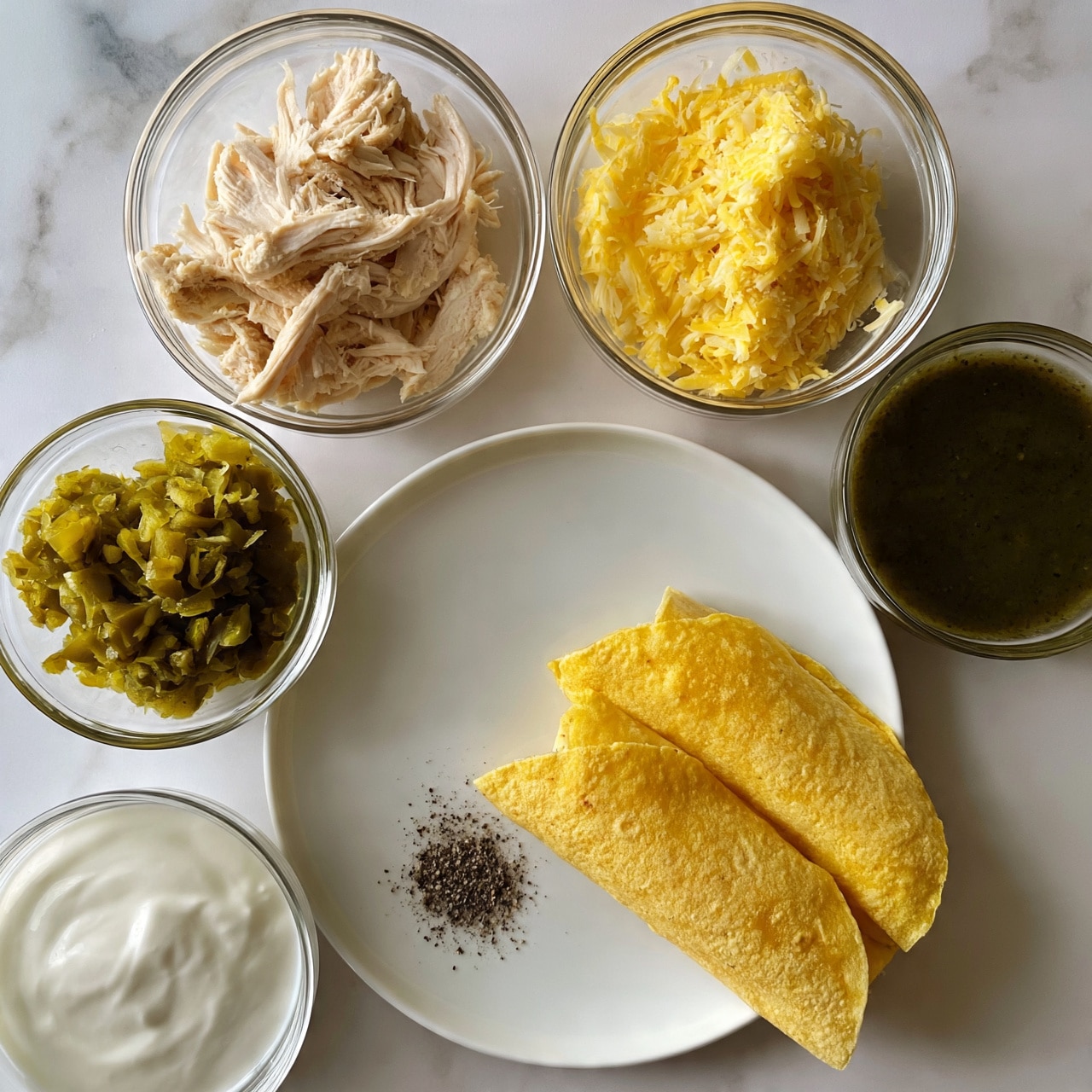 square image A white plate on a white marbled surface holds two folded yellow corn tortillas in the lower right. Surrounding the plate are four clear glass bowls: the top left bowl has shredded cooked chicken in pale beige, the top right bowl contains shredded yellow cheese, the bottom left bowl holds green chopped pickled jalapeños, and the bottom right bowl has a dark green sauce. Below the bowls and to the left of the plate is a small bowl of white sour cream. A small pile of black pepper rests on the white plate near the tortillas. Photo taken with an iphone --ar 4:5 --v 7square image