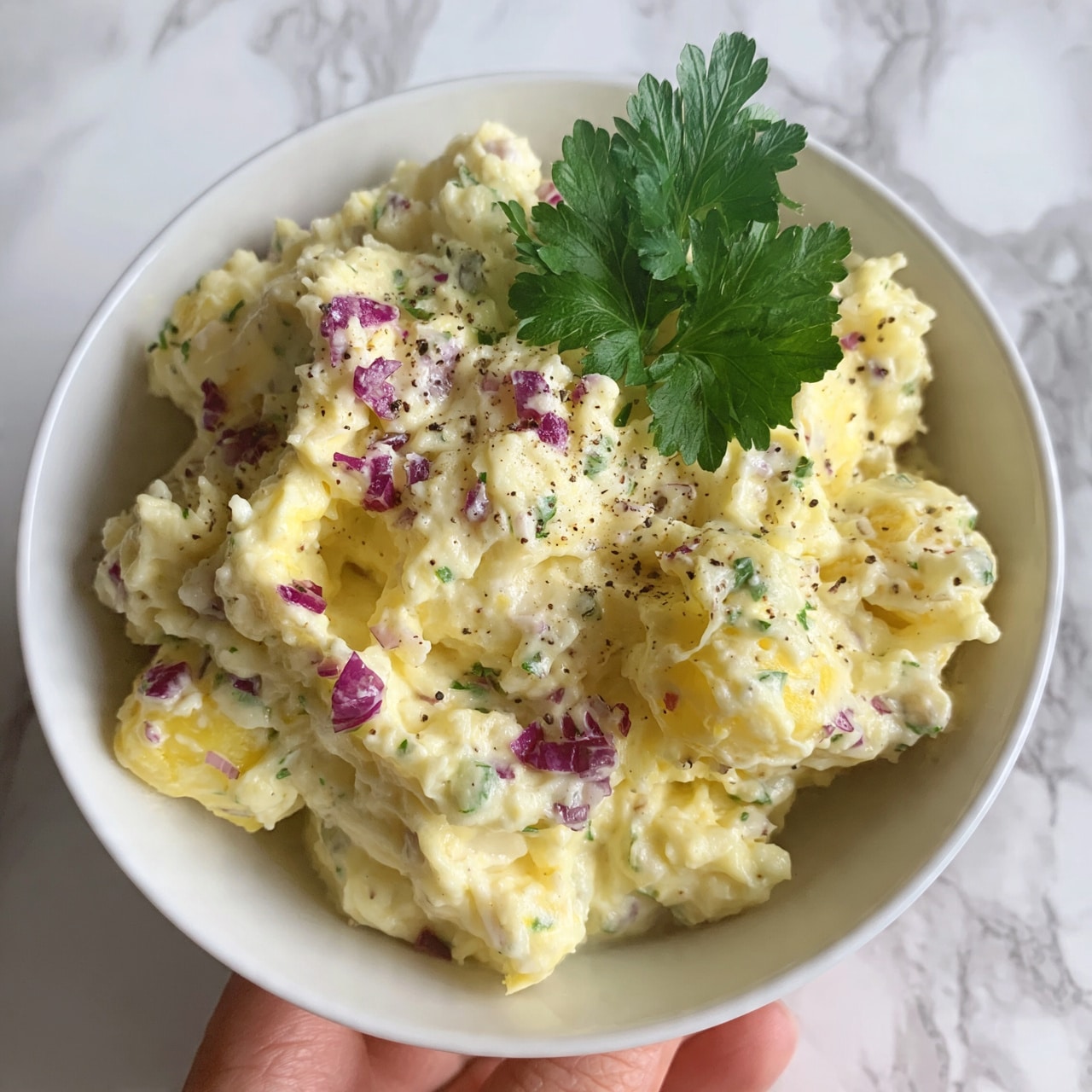 square image A white bowl filled with a creamy light yellow mashed potato salad that has chunks mixed in, topped with small pieces of purple onion, specks of green herbs, and black pepper scattered throughout. There is one green parsley leaf placed on top as a garnish. The white bowl is being held by a person's fingers at the bottom edge, and the background has a white marbled texture. photo taken with an iphone --ar 4:5 --v 7
