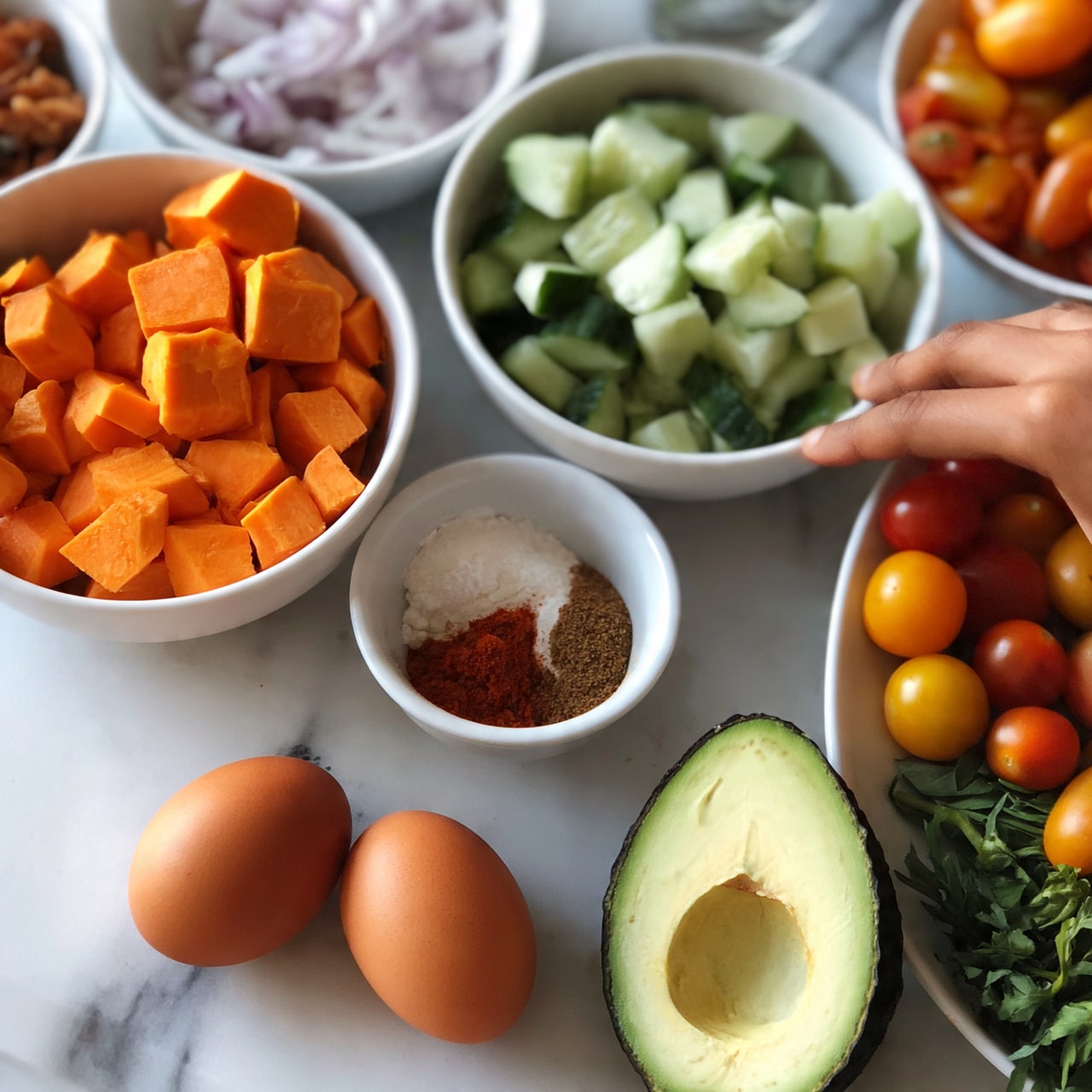 square image The image shows several white bowls on a white marbled surface, each filled with different fresh ingredients. One bowl contains bright orange, cubed sweet potatoes, another has chopped cucumber pieces that are pale green with darker green skin, and a smaller bowl holds finely chopped light purple onions. There is also a small white bowl with three spices arranged separately, showing powdered white, red, and brown colors. In the foreground, two brown eggs rest on the surface near a halved avocado with a dark green outer skin and light green inside with a pit. Cherry tomatoes in red, yellow, and orange tones fill a white bowl, and some fresh green herbs lay next to it. A woman's hand is reaching towards one of the bowls. Photo taken with an iphone --ar 4:5 --v 7