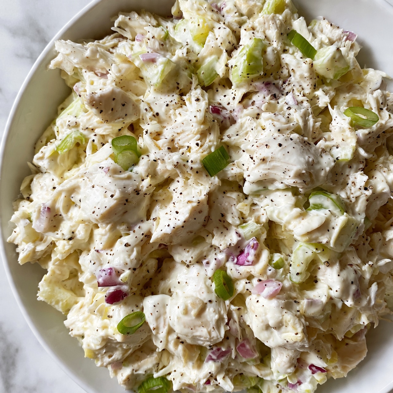 square image A close-up view of a creamy chicken salad served in a white bowl, placed on a white marbled surface. The salad has a mix of shredded white chicken coated in a thick white mayo-like dressing. Small chopped pieces of light green celery, tiny bits of purple-red onion, and slices of green scallion are spread evenly throughout the salad. The salad surface is sprinkled with small black pepper specks, adding texture and contrast to the mostly white and pale pale yellow mixture. photo taken with an iphone --ar 4:5 --v 7