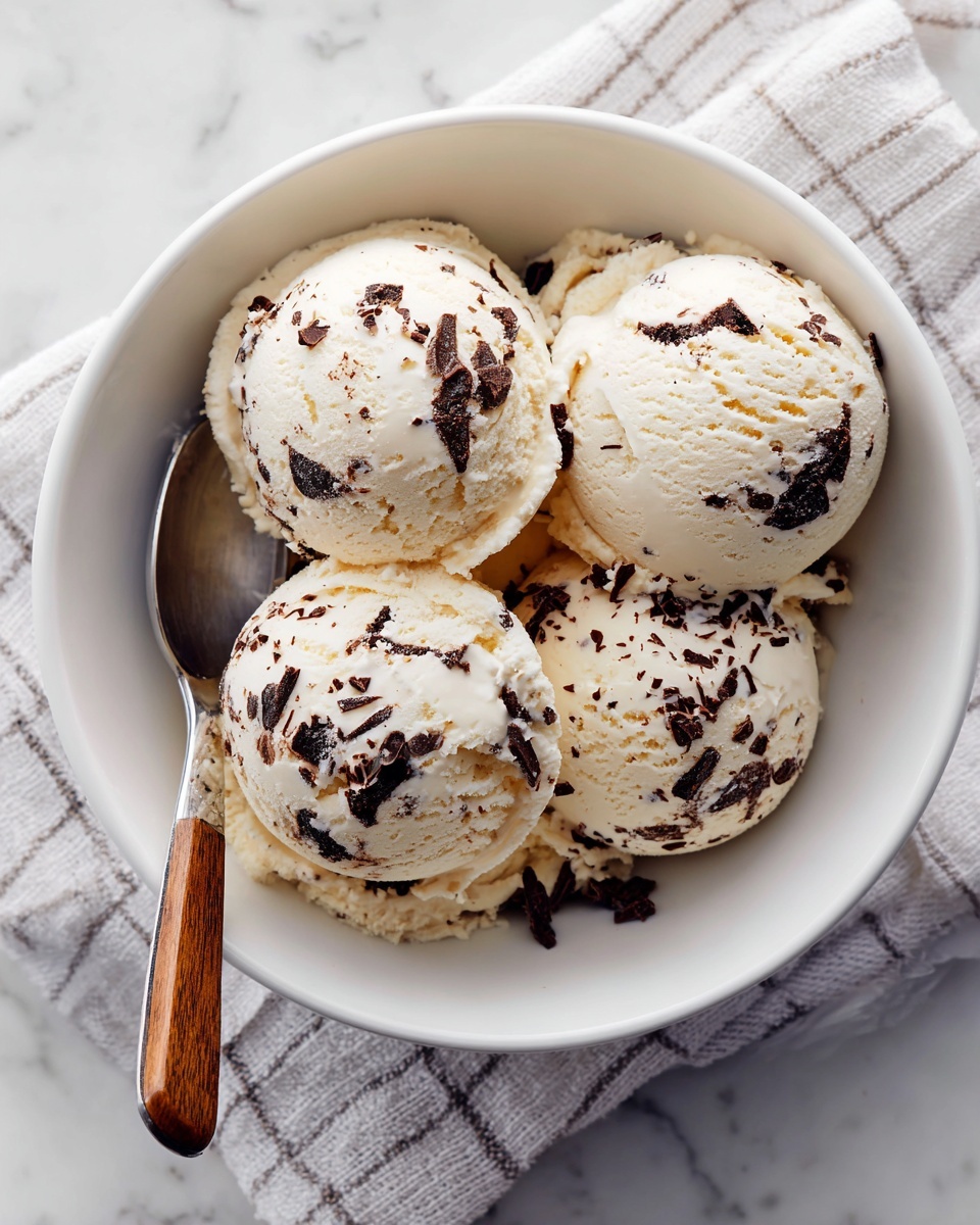 square image A white bowl filled with five round scoops of creamy light beige ice cream, each scoop showing visible dark chocolate pieces mixed inside. The ice cream texture looks smooth and soft with slightly uneven surfaces. A spoon with a silver head and wooden handle rests inside the bowl, partially under the scoops. The bowl is placed on a white marbled surface with a light striped cloth partially under it. photo taken with an iphone --ar 4:5 --v 7