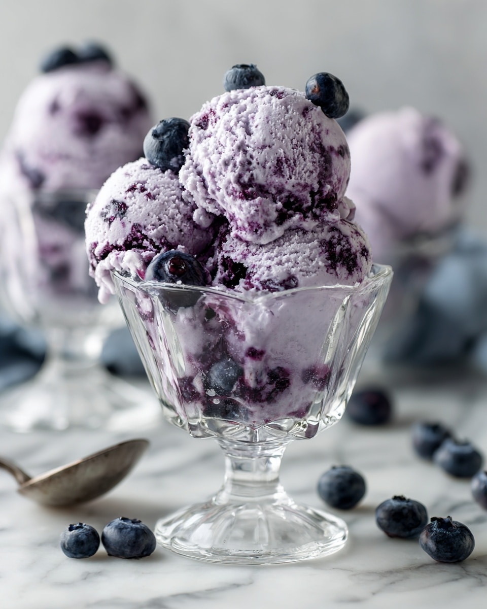square image A clear glass dessert cup filled with three scoops of light purple blueberry ice cream, showing a textured mix of creamy and small blueberry pieces throughout. On top and around the ice cream, several whole dark blue blueberries are placed as garnish. The cup is set on a white marbled surface, with a few scattered blueberries nearby. In the background, there is another similar glass cup with the same ice cream, softly out of focus. The light is soft and natural, highlighting the creamy texture and fresh berries. Photo taken with an iphone --ar 4:5 --v 7