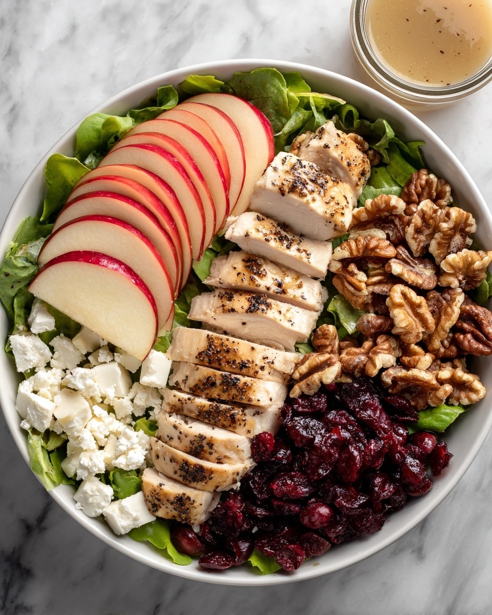 A white bowl filled with a base of green salad leaves. On top, there are three main sections: the center holds sliced cooked chicken, light brown with some black pepper seasoning, arranged in a neat row. To the left, there is a fan of thin red apple slices with white inner flesh, partially under the chicken. On the right, there are large walnut pieces that look crunchy, next to a pile of dark red dried cranberries. Small white cheese crumbles are placed at the bottom left of the salad leaves under the chicken. In the upper right corner, there is a small clear glass jar with light brown dressing. Everything is on a white marbled texture surface. photo taken with an iphone --ar 4:5 --v 7
