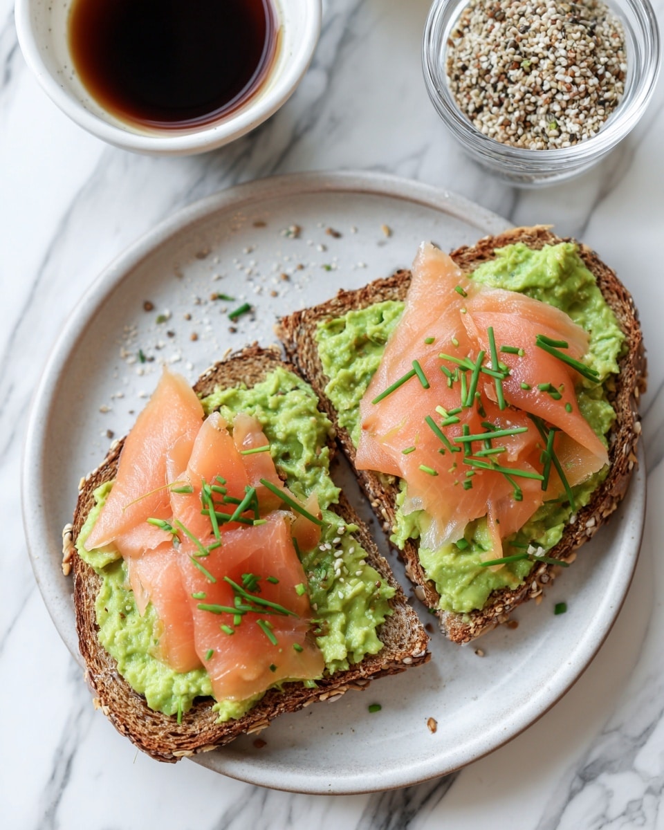 Two slices of multigrain toast lie on a white plate with a white marbled background. Each slice has a green mashed avocado layer spread evenly on top. One slice is topped with thin, folded light pink smoked salmon, while small green chive pieces are sprinkled over the salmon. The other slice shows just the avocado spread in the background. To the right of the plate, there is a small clear bowl of mixed seeds, and on the left side, a small bowl of dark soy sauce is partially visible. Photo taken with an iphone --ar 4:5 --v 7