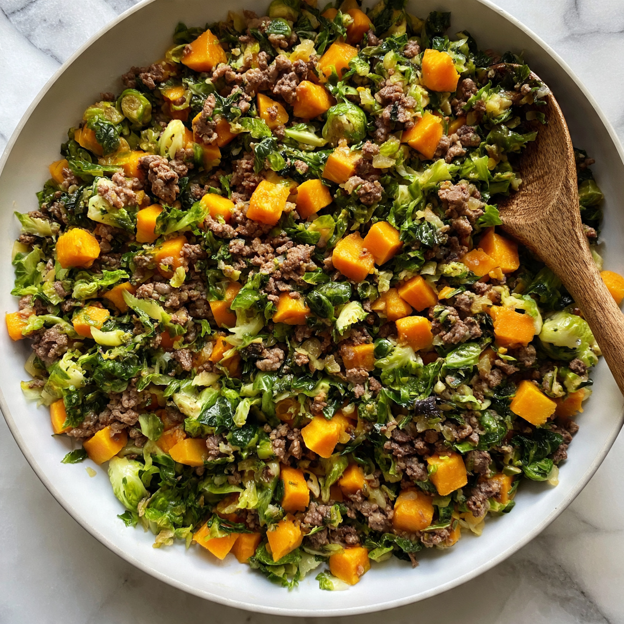 square image The image shows a white pan filled with cooked ground beef mixed with bright orange cubes of sweet potato and chopped green Brussels sprouts. A wooden spoon rests inside the pan on the right side, partly covered with some of the dish. The vegetables and meat are mixed evenly, creating a colorful and textured look with the green, orange, and brown tones all visible. The pan sits on a white marbled surface. Photo taken with an iphone --ar 4:5 --v 7