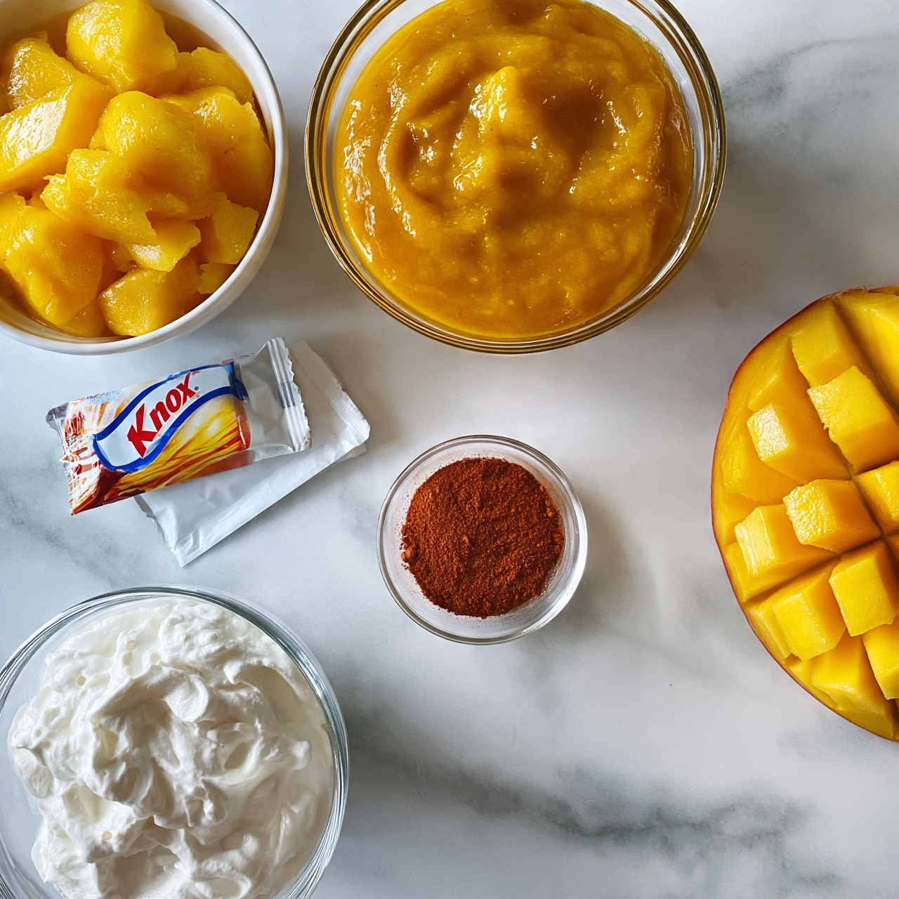 square image The image shows six items on a white marbled surface arranged neatly. At the top left, there is a white bowl full of bright yellow mango chunks. To the right, there is a clear glass bowl filled with thick, smooth mango puree that has a rich golden-yellow color. Below the mango chunks, there is a Knox gelatin package with one envelope lying flat beside it. At the center, there is a small clear bowl filled with reddish-brown spice powder. On the bottom left corner, there is a clear glass bowl filled with creamy white whipped cream. Finally, on the right edge, there is a fresh mango sliced into large cubes but still attached to the skin. Photo taken with an iphone --ar 4:5 --v 7