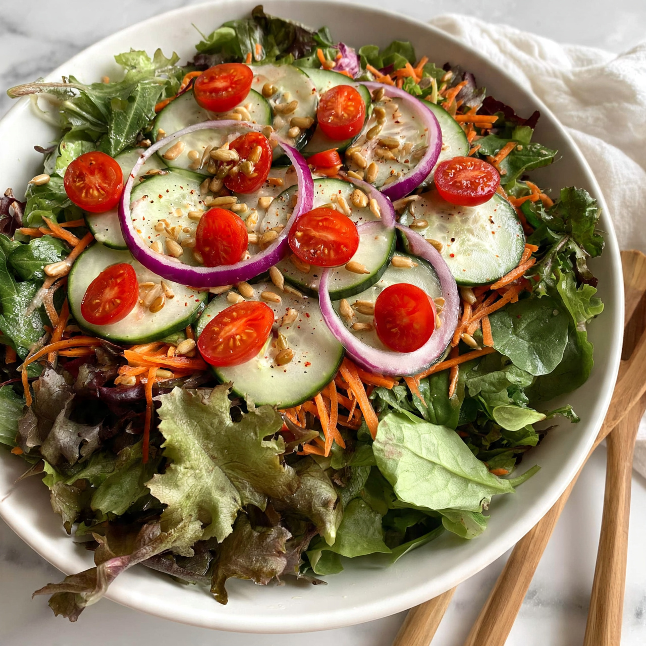 square image A white bowl filled with a fresh salad consisting of two layers: the bottom layer is a mix of leafy greens in various shades of green with rough and smooth textures, and the top layer is topped with thin slices of cucumber, halved cherry tomatoes in bright red, thin rings of purple onion, shredded orange carrot, and sprinkled lightly with small tan sunflower seeds. The bowl is set on a white marbled surface with a soft cloth nearby, and wooden salad utensils are placed close to the bowl. photo taken with an iphone --ar 4:5 --v 7