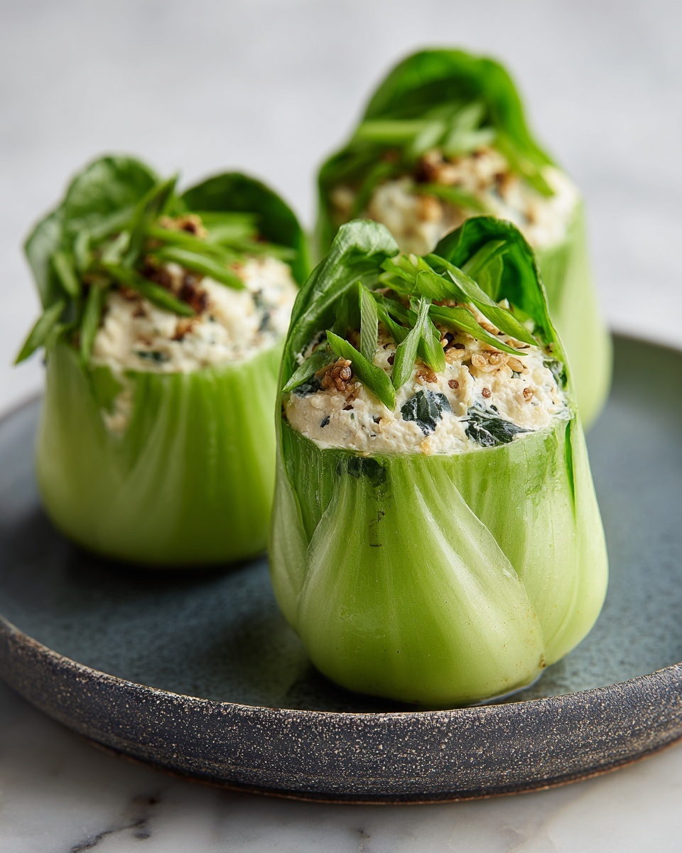 This image shows three green bok choy pieces standing upright on a dark gray round plate with a rough texture, placed on a white marbled surface. Each bok choy is hollowed out and filled with a creamy white mixture that has small brown bits and green herbs mixed inside. The bok choy leaves are crisp and vibrant green, slightly open at the top to hold the filling, with the stems tightly closed at the bottom. The focus is on the front bok choy, showing clear details of the creamy filling and fresh green leaves. Photo taken with an iphone --ar 4:5 --v 7