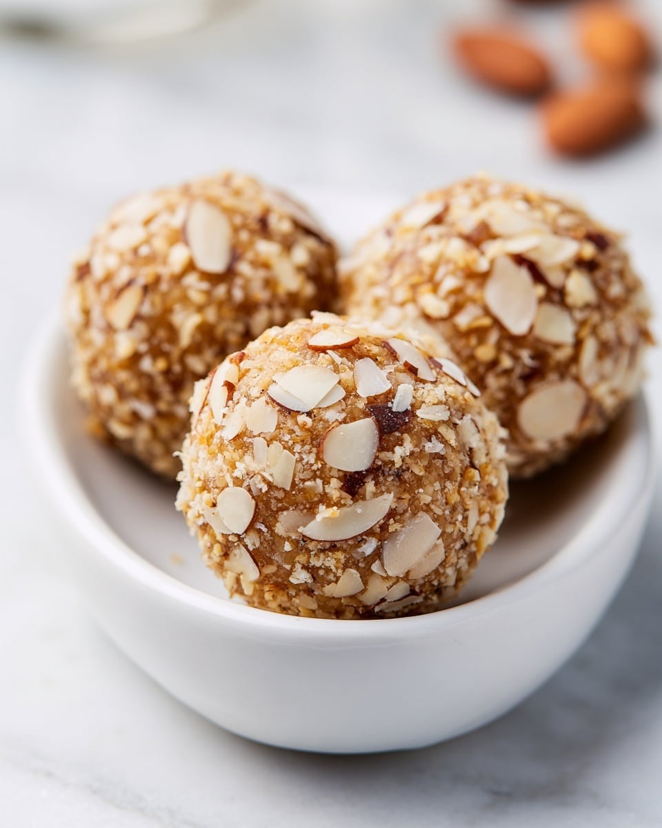 The image shows three round snack balls covered with thin, light brown almond slices. They are placed in a white oval bowl with a smooth texture. The balls have a rough texture from the almond slices, and each ball is evenly coated with them. The bowl is set on a white marbled surface, giving a clean and bright background. The focus is on the middle snack ball, with the other two slightly blurred in the distance. photo taken with an iphone --ar 4:5 --v 7