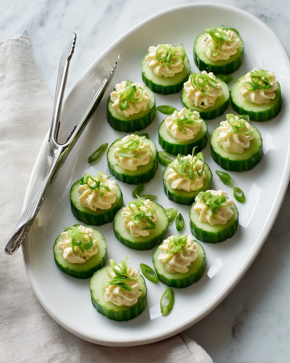 A white oval plate holds 17 round cucumber slices arranged in neat rows. Each cucumber slice has a light green smooth top layer of creamy beige spread dolloped generously, creating a soft texture. On top of each dollop, small bright green thin round slices of scallions add a fresh contrast. Stainless steel tongs rest on the left edge of the plate, and a light-colored cloth is visible underneath on the left. The whole scene sits on a white marbled surface. Photo taken with an iphone --ar 4:5 --v 7