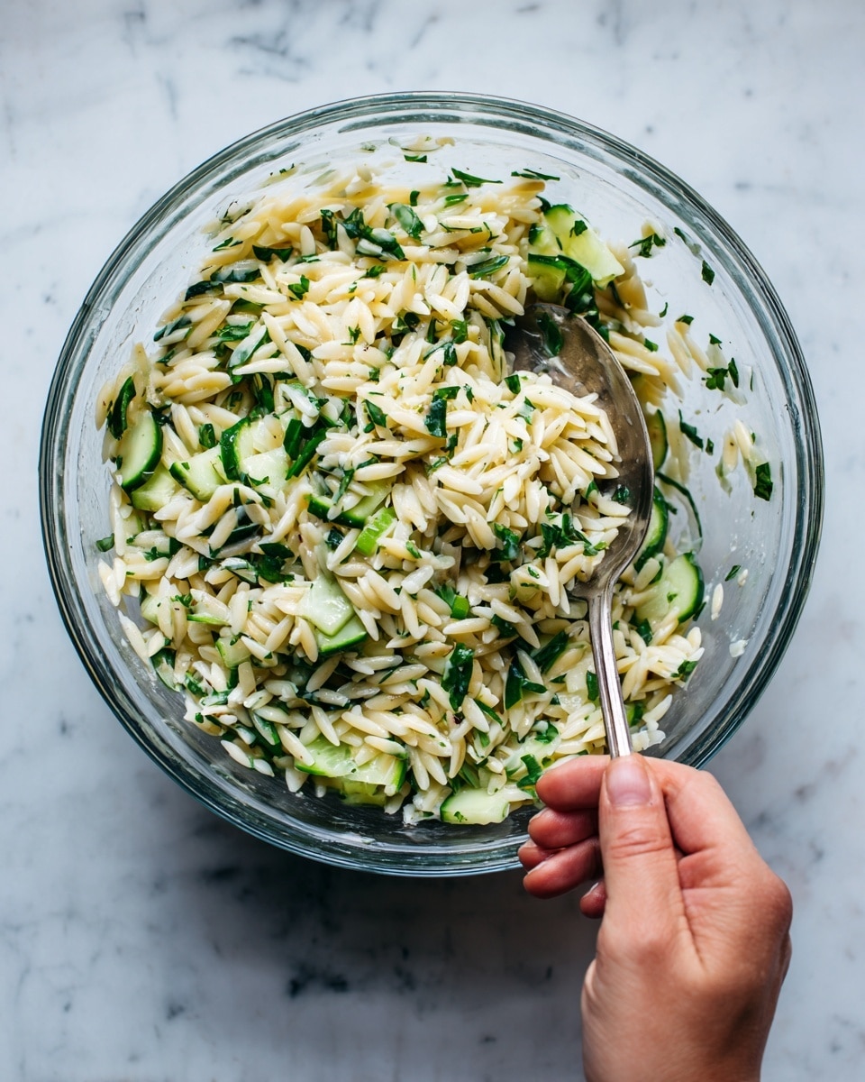 A clear glass bowl filled with a fresh orzo salad that has small, rice-shaped pasta mixed with chopped cucumber pieces and bits of green herbs spread evenly throughout. The salad has a light, creamy white color from the orzo, bright green from the cucumber slices, and specks of deeper green from the herbs. A woman's hand is holding a large metal spoon inside the bowl, ready to mix or serve the dish. The bowl is placed on a white marbled surface that contrasts softly with the colors of the salad. Photo taken with an iphone --ar 4:5 --v 7
