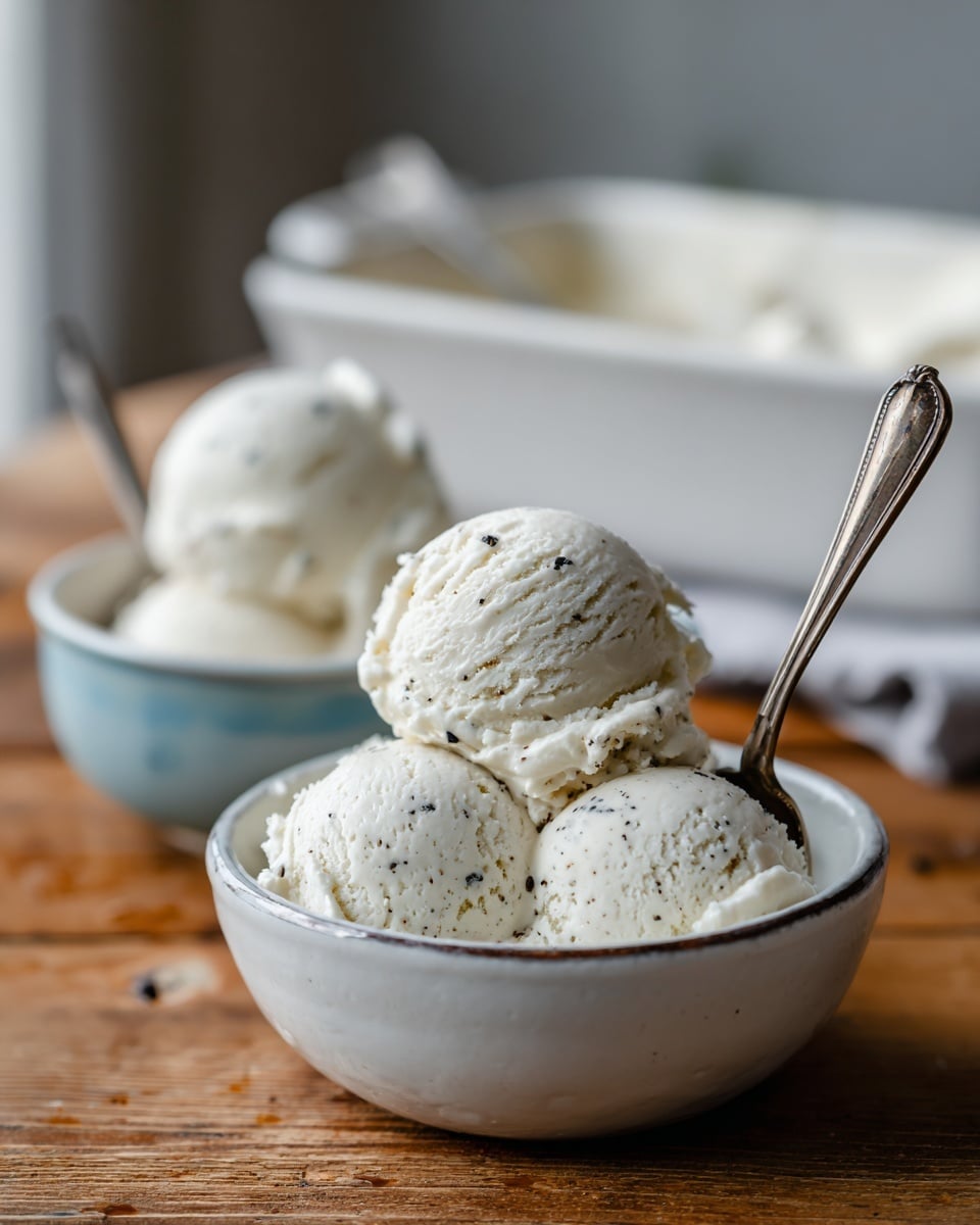 The image shows two scoops of white ice cream with small vanilla bean specks held in two small white bowls that have a blue outside texture. In the front bowl, there are three round scoops stacked, one on top of the other. A vintage silver spoon is placed inside the bowl, leaning forward. In the background, another bowl holds one scoop of ice cream with a similar silver spoon. Behind them, a white baking dish is blurred but visible. The setup is on a wooden surface and the overall look is soft with natural light. photo taken with an iphone --ar 4:5 --v 7