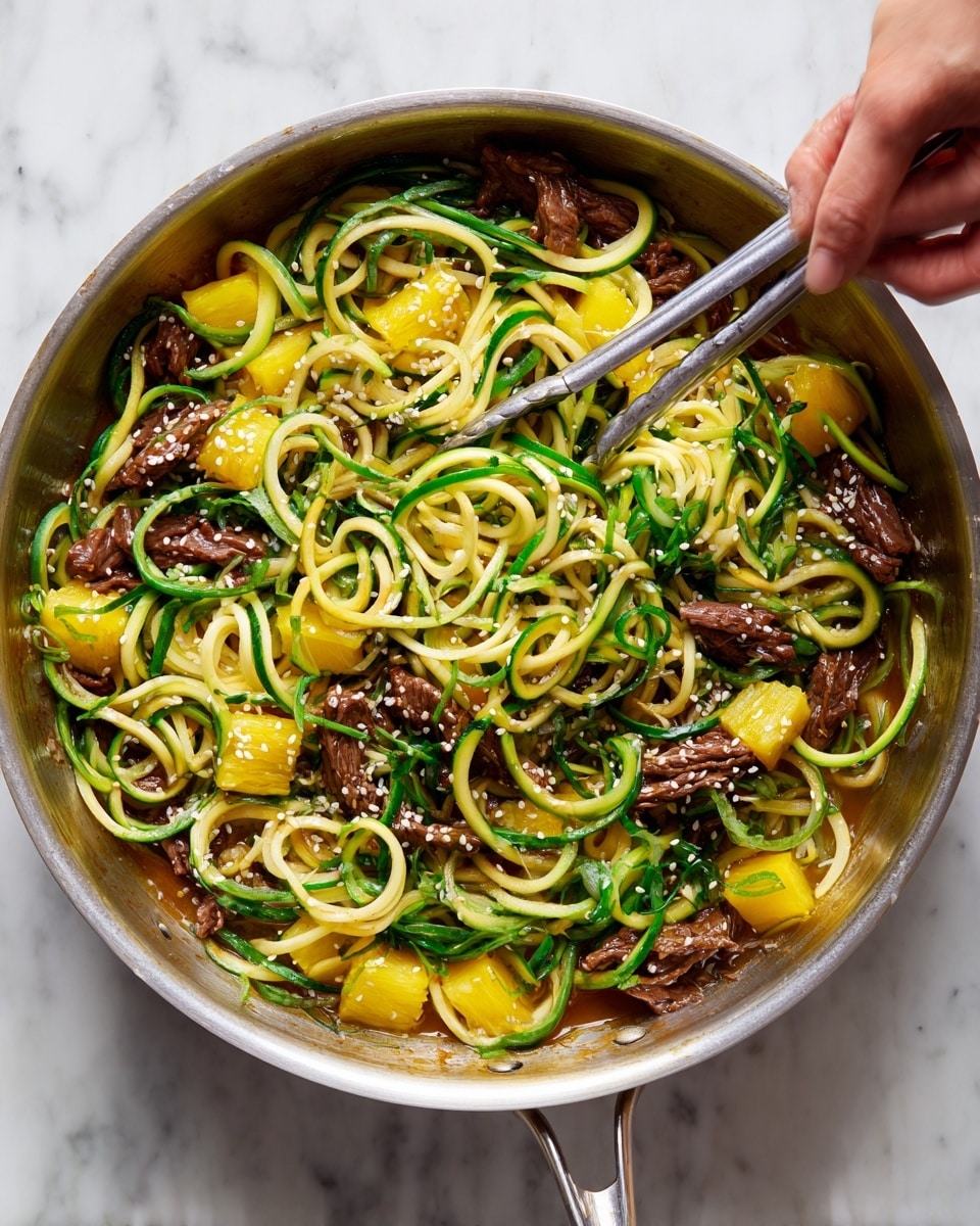 A large round silver pan filled with spiral-shaped light green and pale yellow zucchini noodles mixed with thin dark brown strips of beef and bright yellow chunks of pineapple, all coated in a slightly shiny brown sauce. White sesame seeds are sprinkled on top, adding a delicate texture. A woman’s hand holding silver tongs is picking up some of the food from the right side of the pan. The pan sits on a white marbled surface. photo taken with an iphone --ar 4:5 --v 7