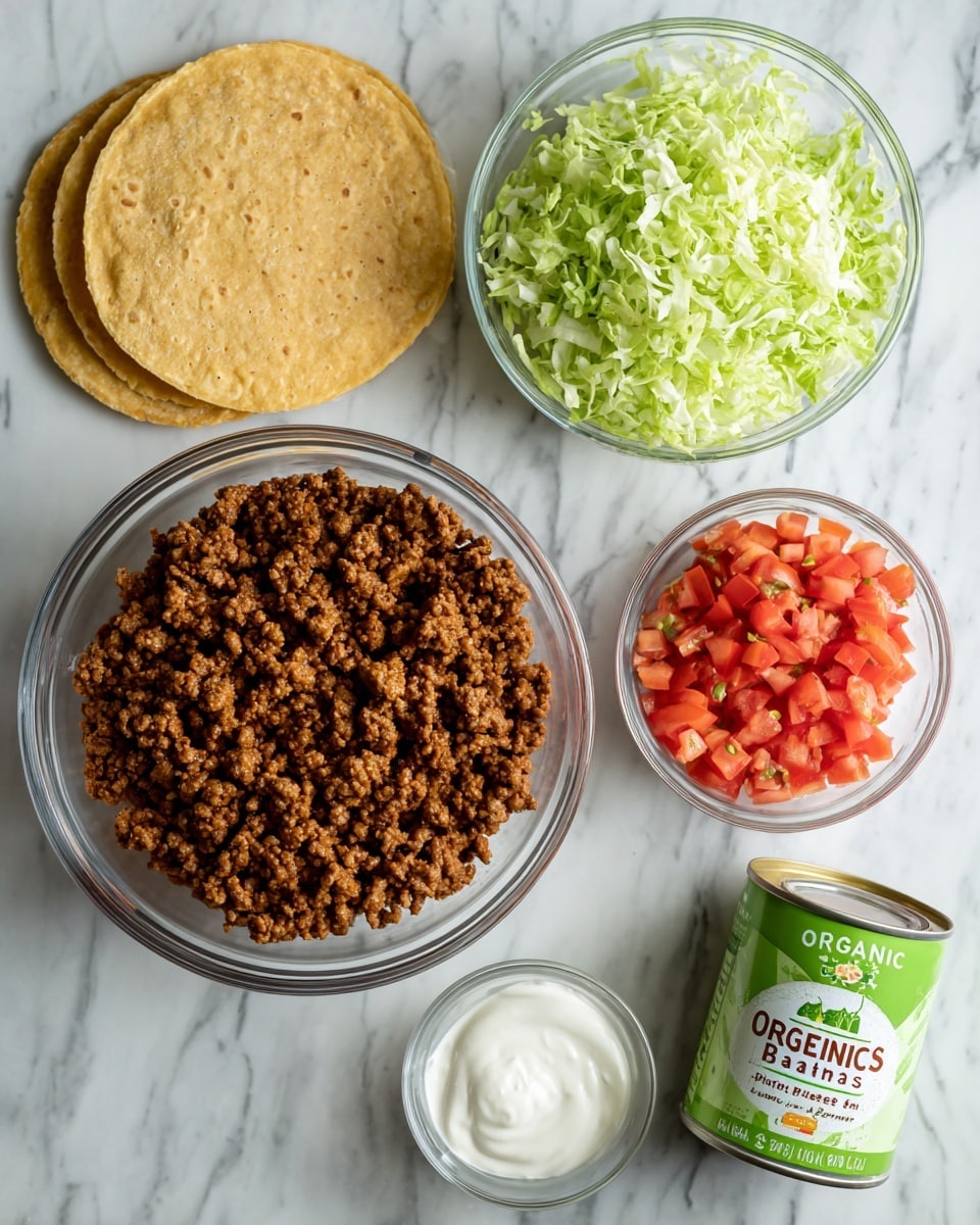 The image shows ingredients for a taco dish arranged on a white marbled surface. In the center, there is a clear glass bowl filled with cooked brown ground meat. Above it, a small clear bowl holds shredded light green lettuce, and to the right of the lettuce, another small clear bowl contains diced red tomatoes. On the right side of the meat bowl, there is a green and white can labeled